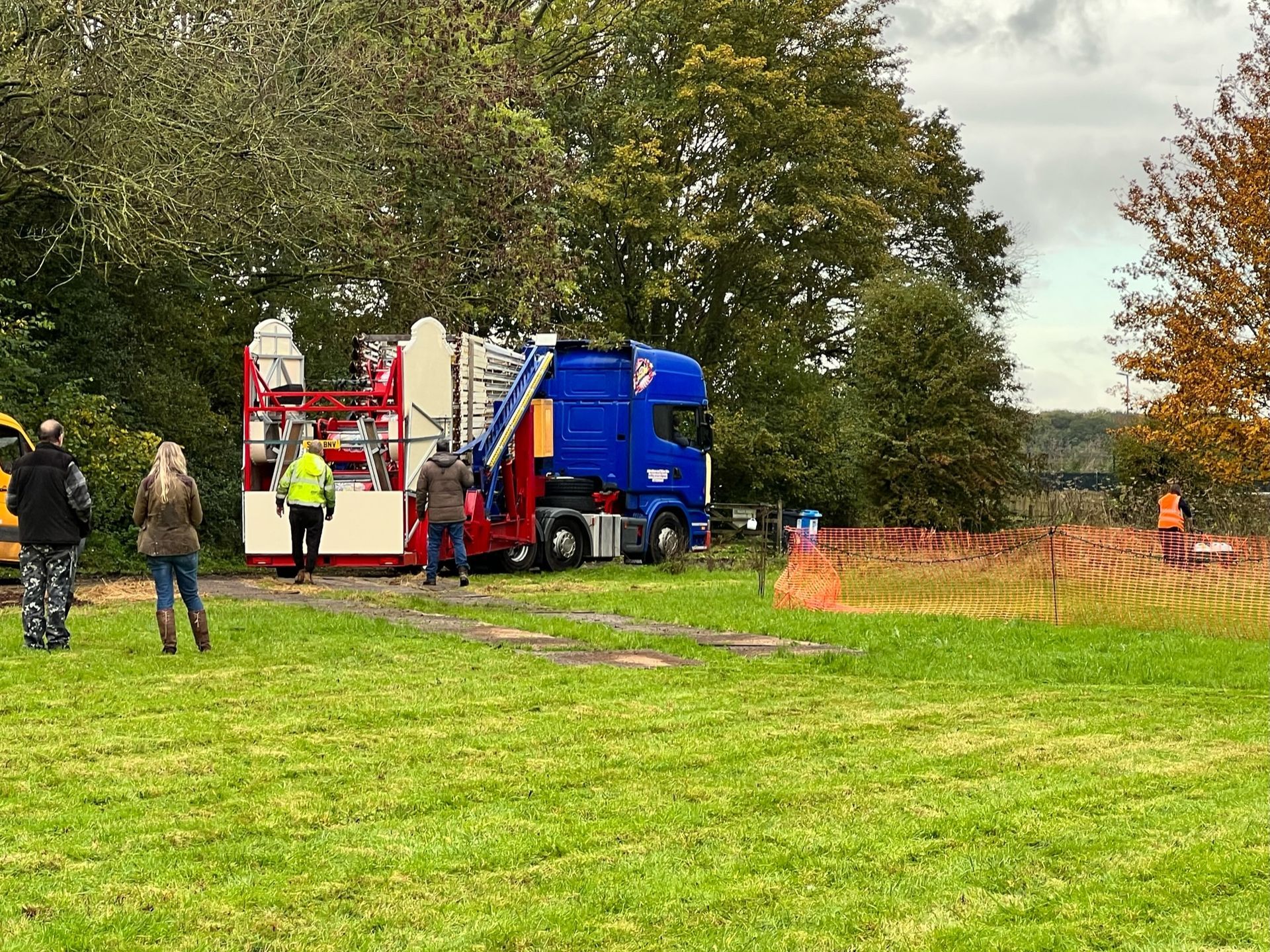 A blue semi-truck with a ride trailer on grass; people watch. Trees and orange safety fencing surround.