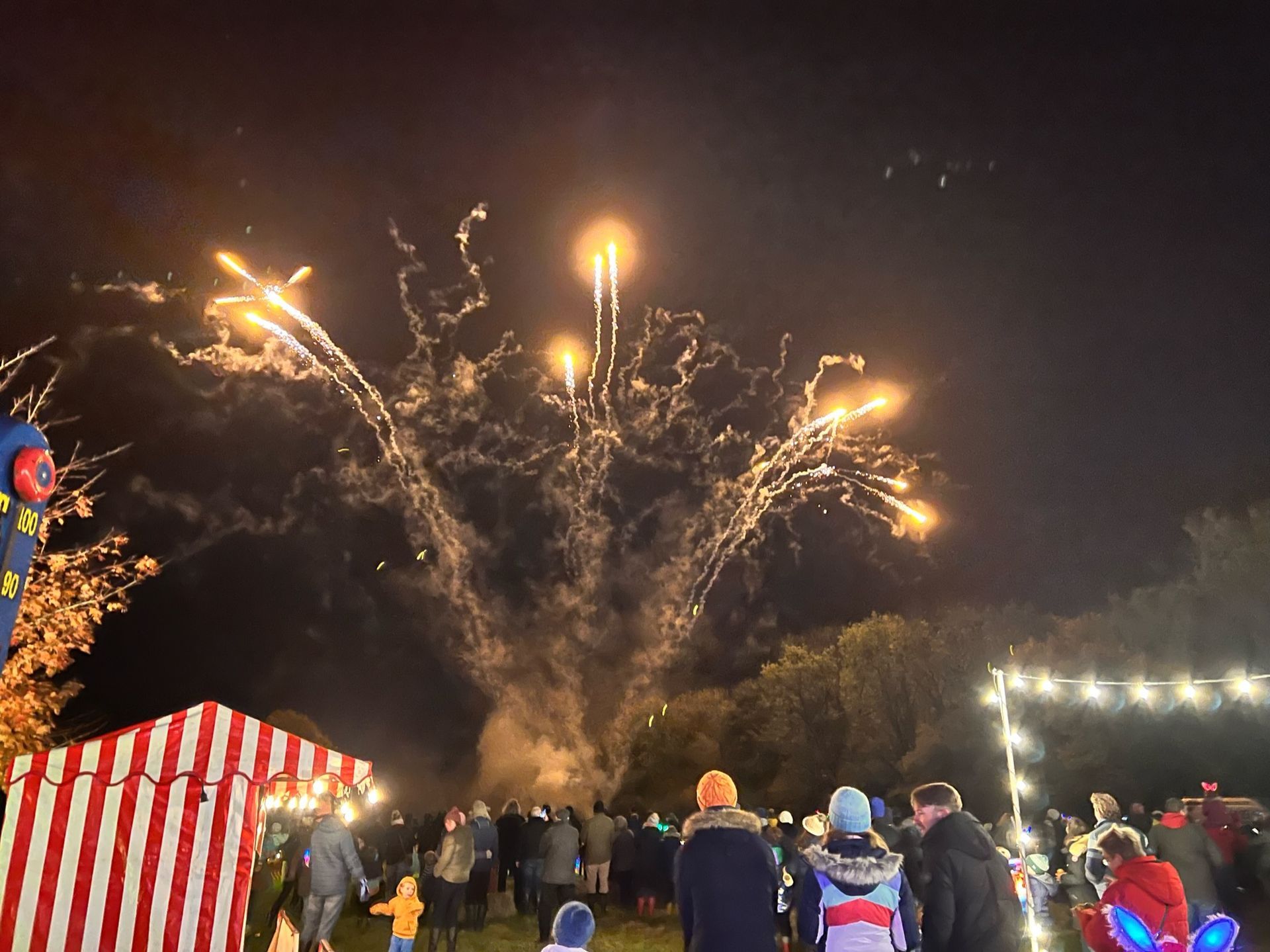 Fireworks bursting over a crowd gathered in a field with a tent and string lights.