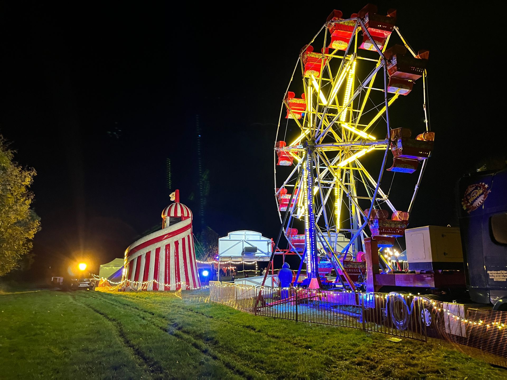 Ferris wheel and carnival game lit up at night on a grassy field.