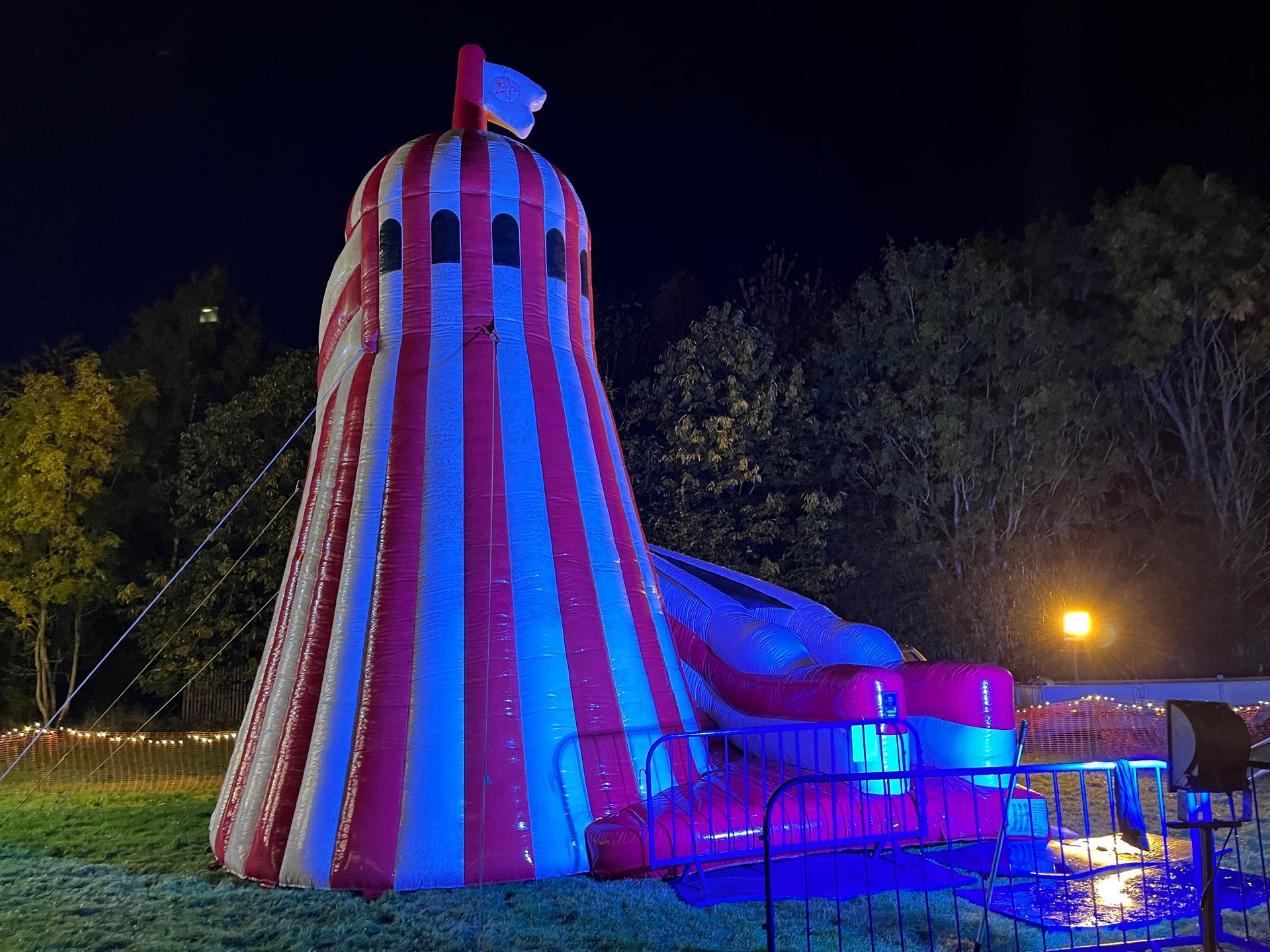 A large inflatable red and white striped slide lit with blue light sits on grass at night.