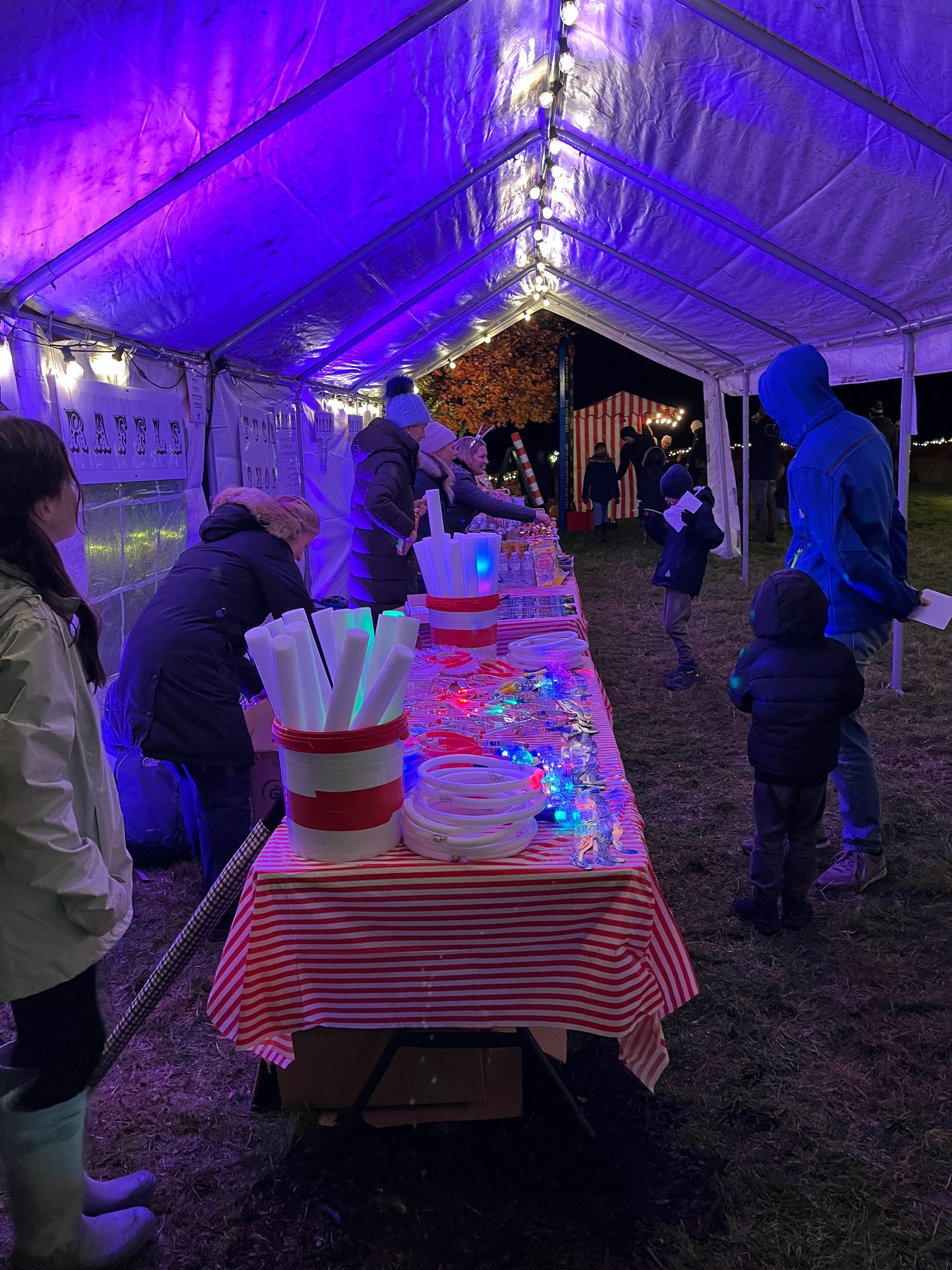 People at a brightly lit snack table under a tent at night. Red and white tablecloth, candy, and treats.