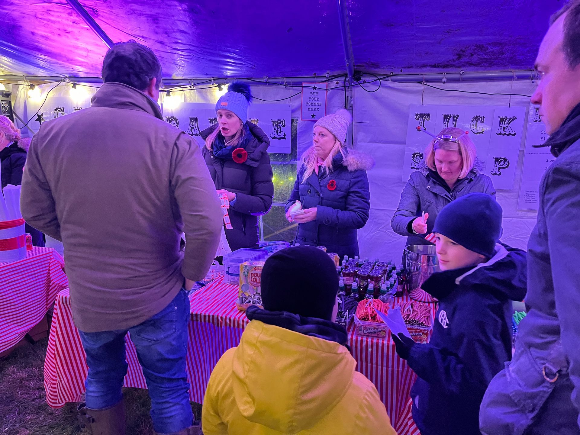 People at a festive outdoor stand, illuminated with purple light, selling items. Some wear poppies, children interact.