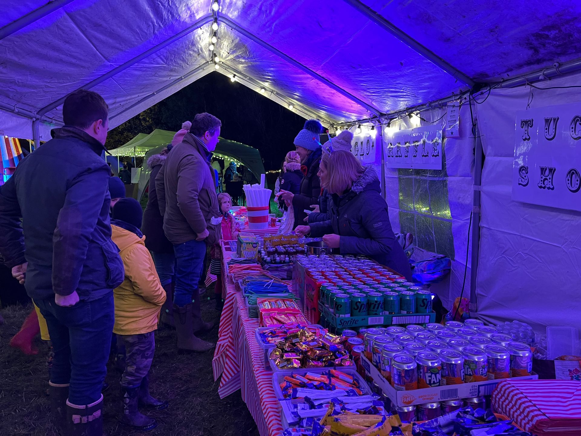 People at a brightly lit concession stand, selling snacks and drinks at an outdoor event.