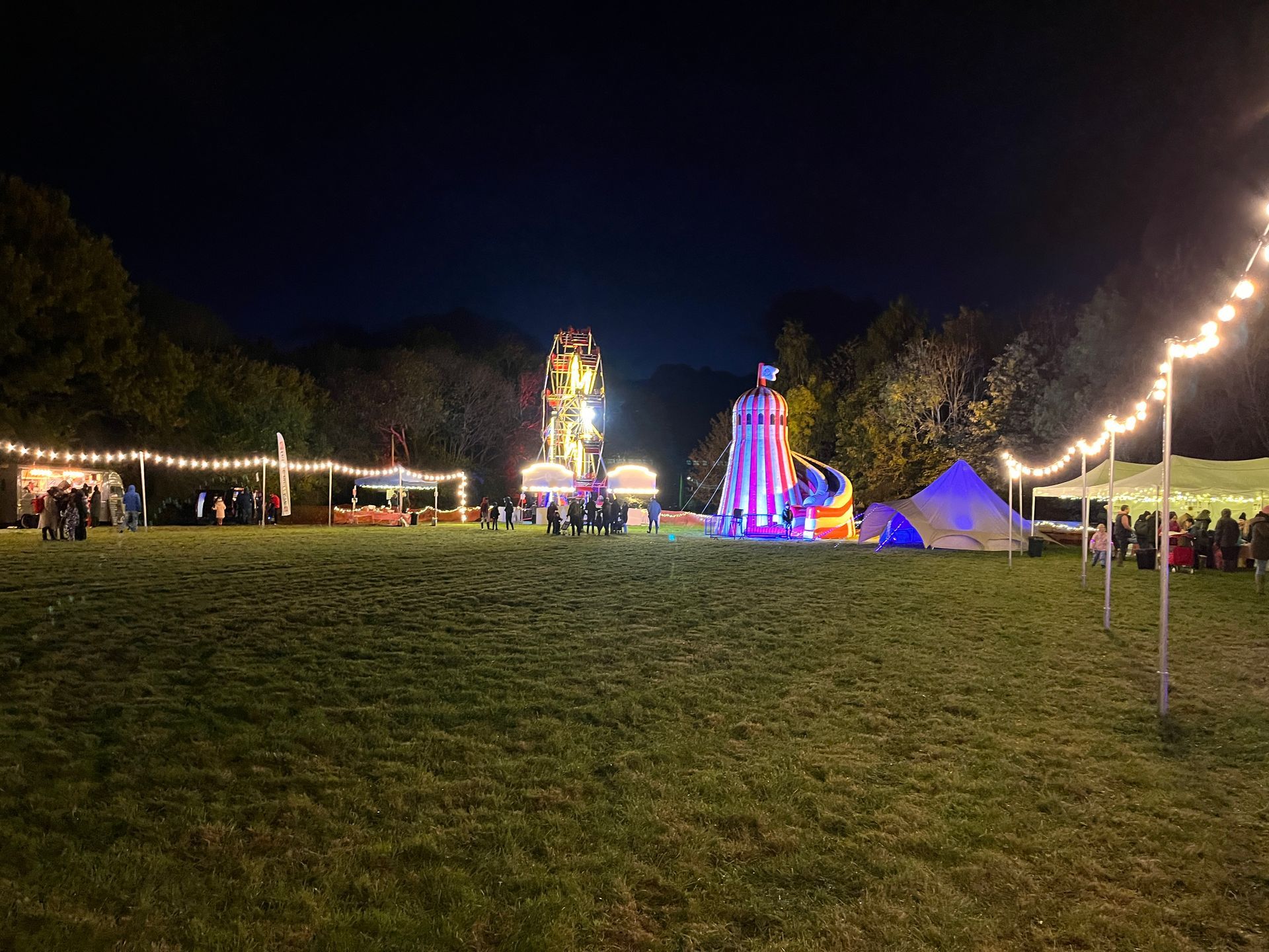 Night fairground with illuminated rides, tents, and string lights in a grassy field.