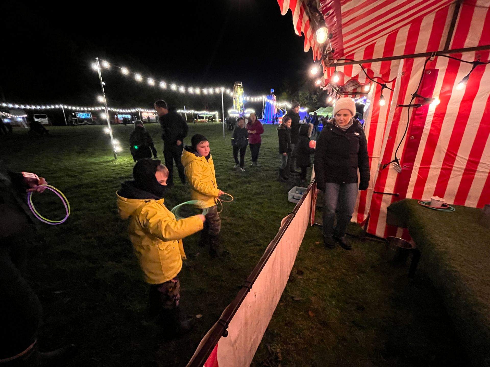 Children playing a ring toss game at a brightly lit outdoor fair at night.