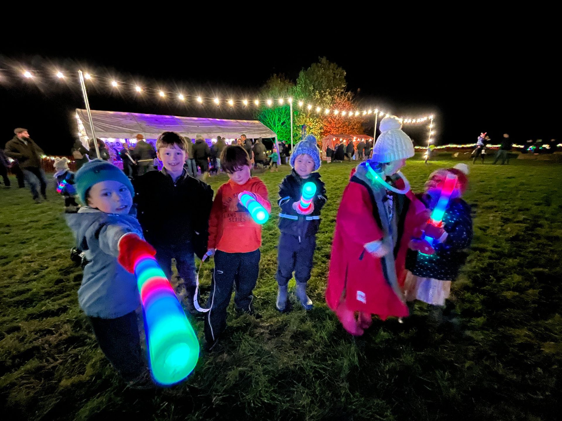 Children holding glowing sticks, smiling at night event with lights and a tent.