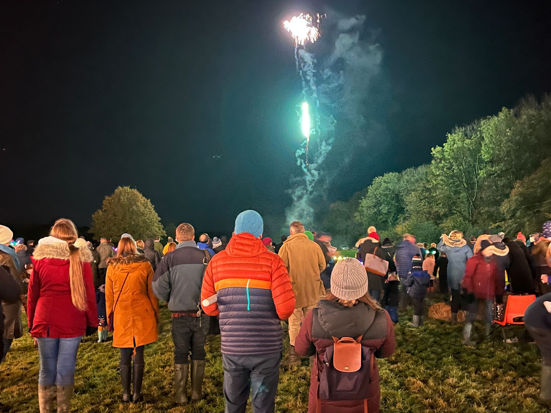 People watching fireworks in a dark field, some with coats and hats, a burst of light in the sky.