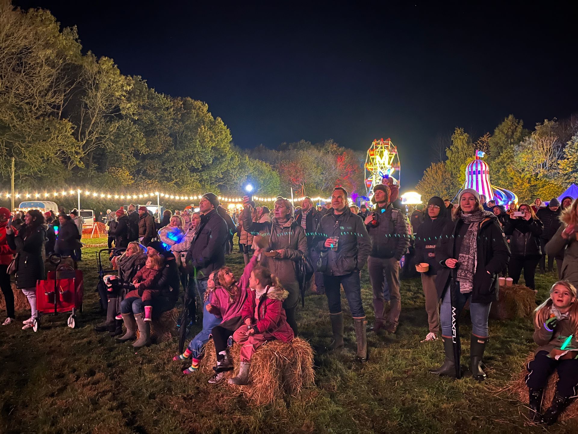 Crowd at an outdoor event at night. People stand, sit on hay bales, with lit attractions in the background.