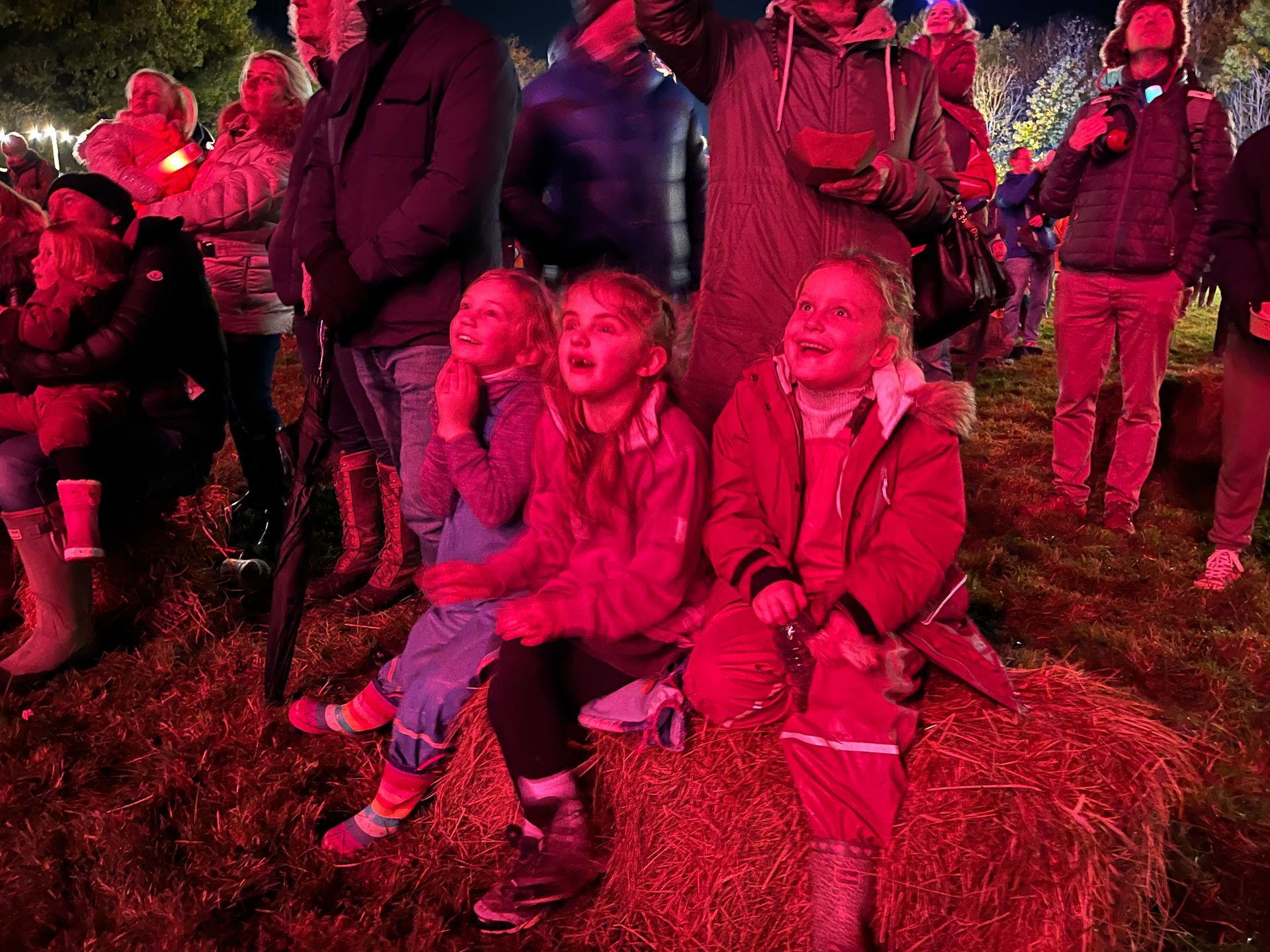 Children sitting on hay bales, illuminated by red light, watching an outdoor event at night.