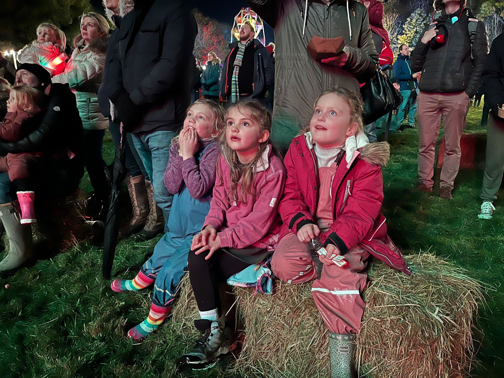 Three girls sit on hay bales, gazing upwards, faces lit. Outdoor event, dark setting.