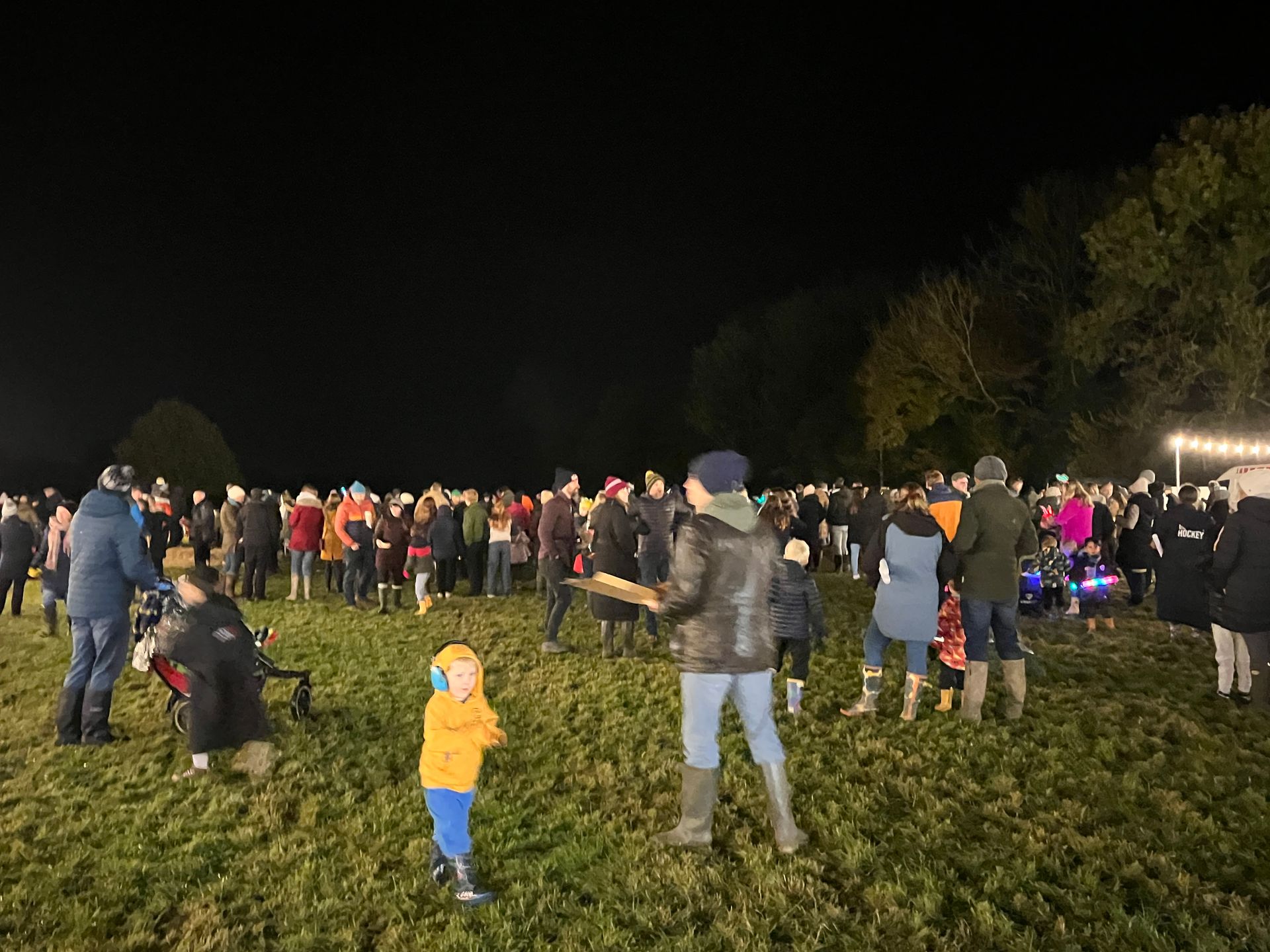 Large crowd at night in a field. People gathered, likely for an event. A small child in a yellow coat stands in front.