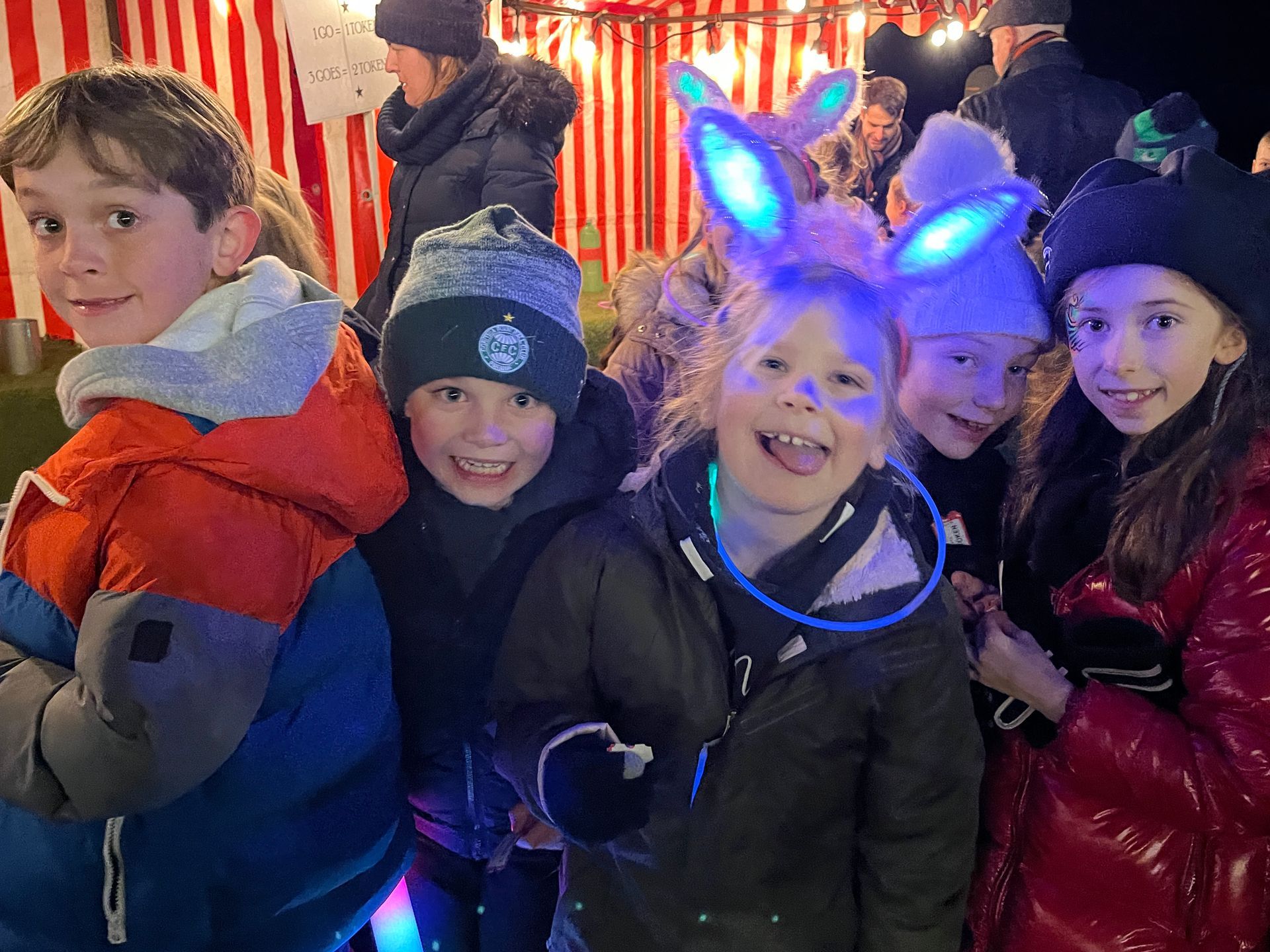 Group of children smiling outdoors at night, festive setting. Some have illuminated accessories.