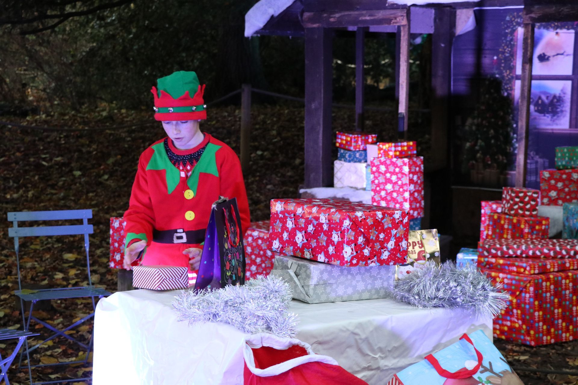 Elf at a holiday display. Red coat, green hat, and wrapped gifts.  Outdoor setting.