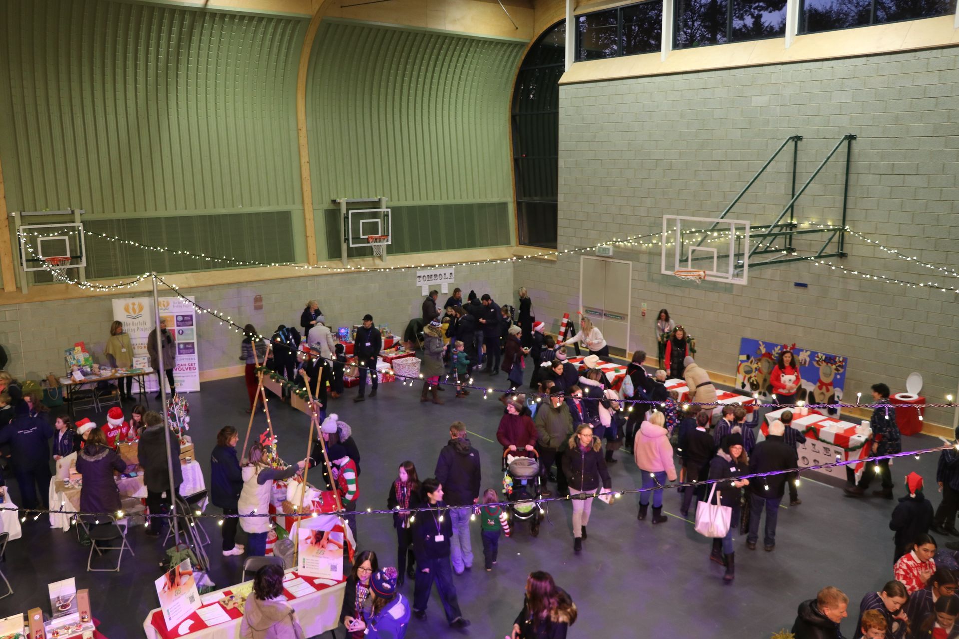 Christmas market in a gymnasium, many people browsing stalls, overhead string lights.