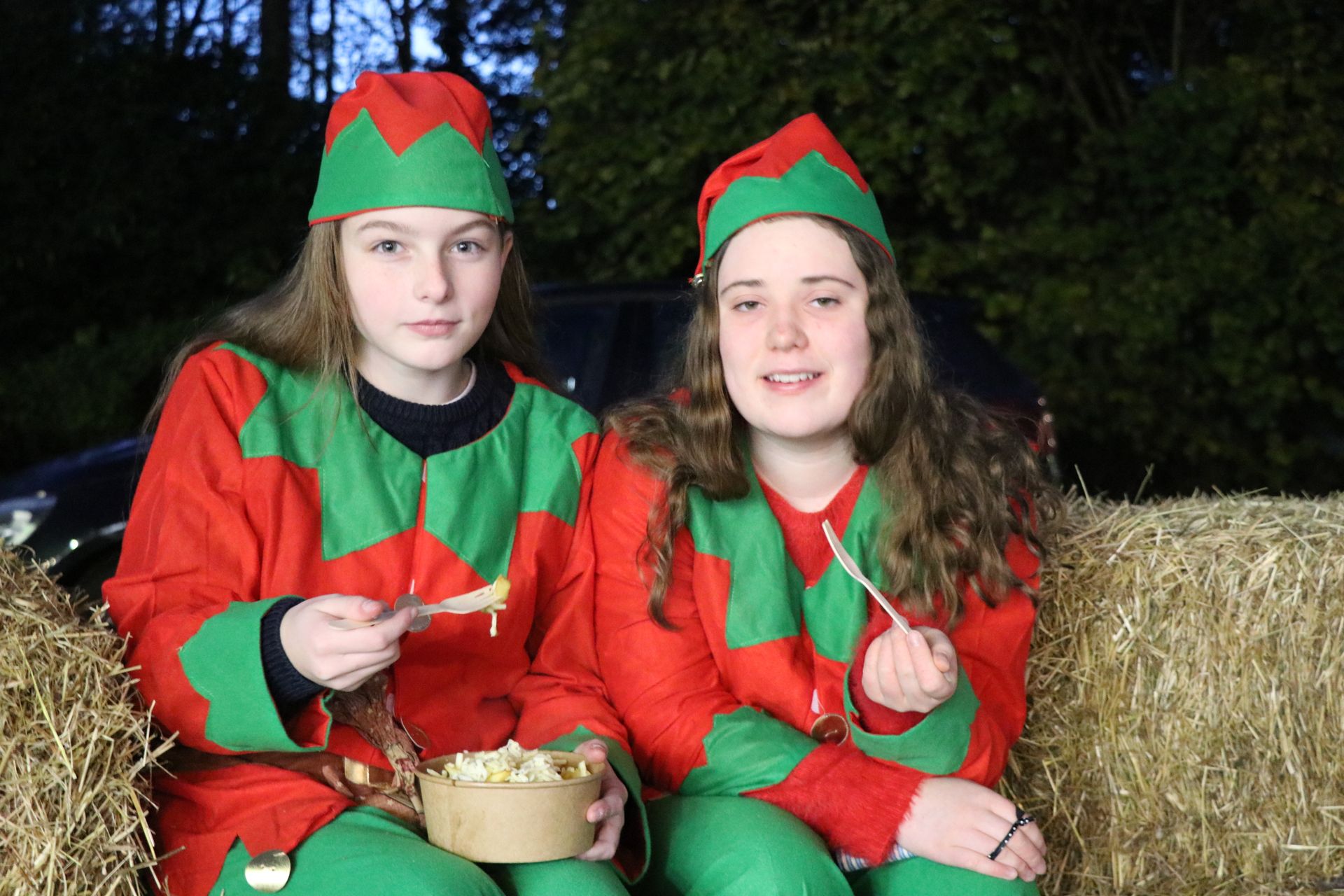 Two people in elf costumes eating from containers, seated on hay bales.