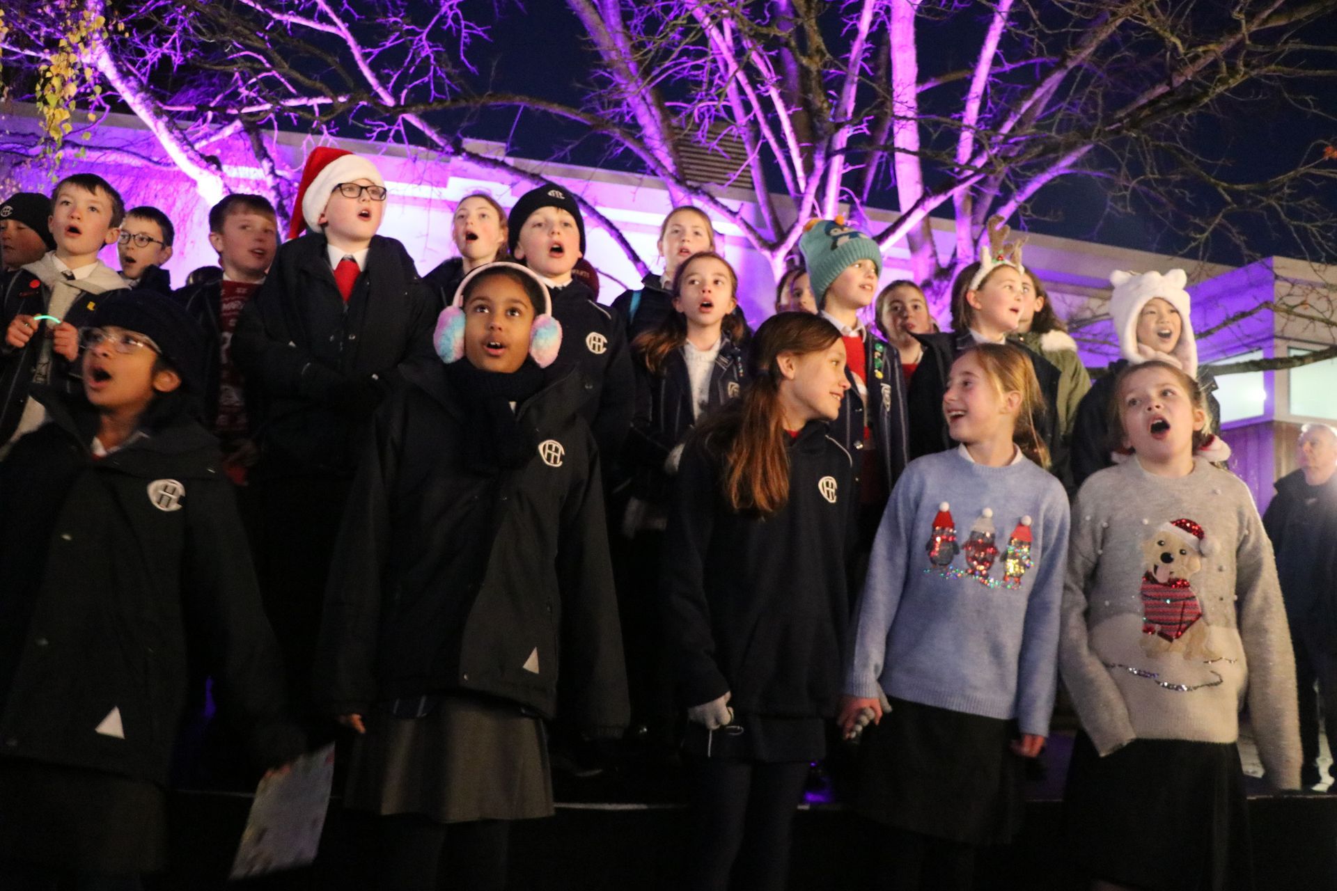 Children singing Christmas carols outdoors at night, illuminated by purple lights.