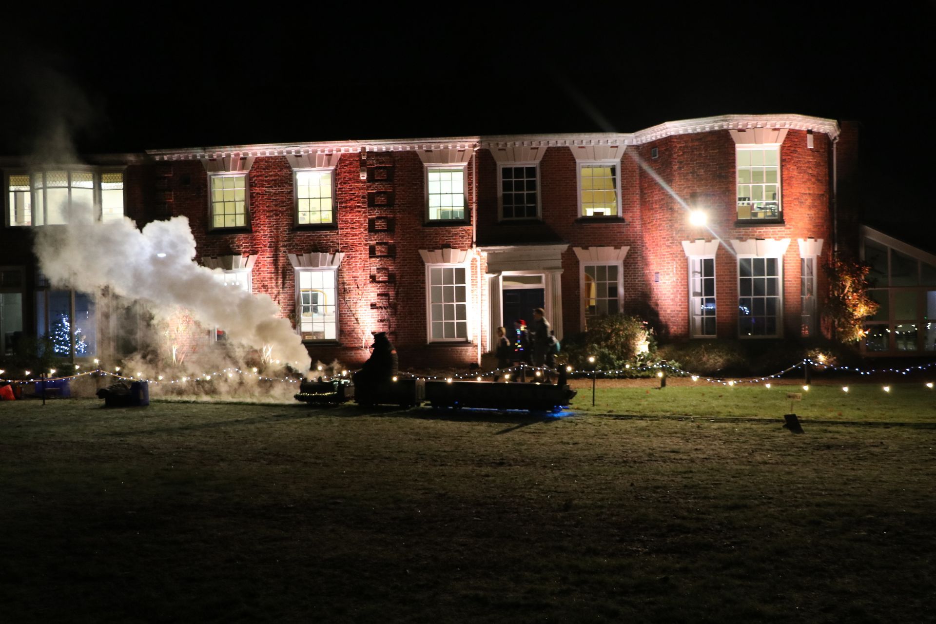 A house lit up at night with a train blowing out smoke on its lawn.