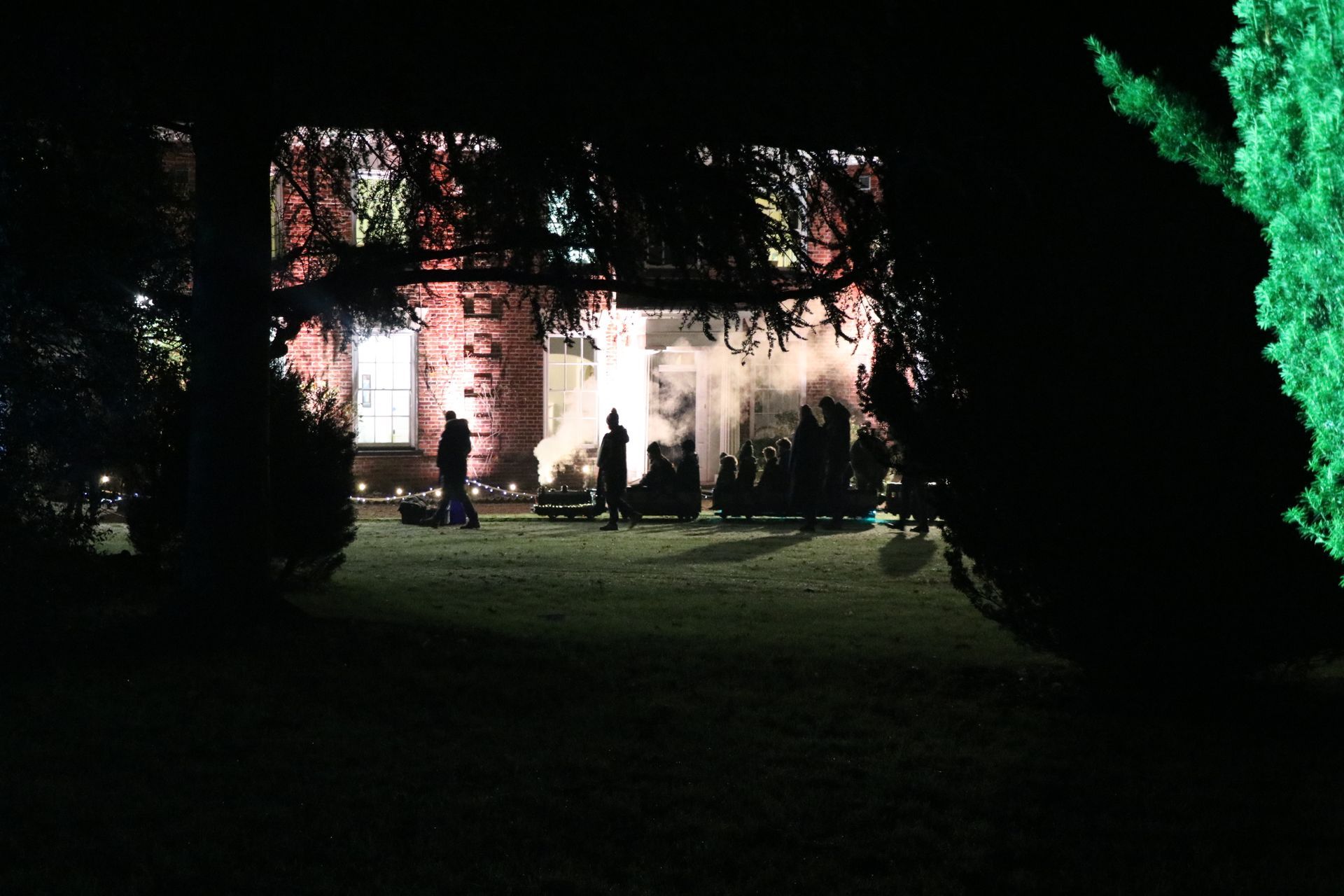 Night scene: Silhouetted figures gather in front of a brightly lit building emitting smoke, framed by trees.