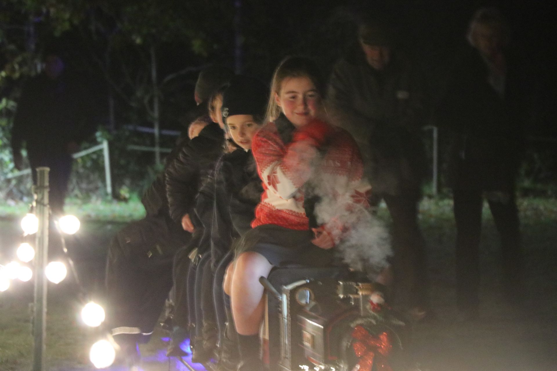 People riding a small train at night, surrounded by lights and fog. One person is holding a fluffy, white object.