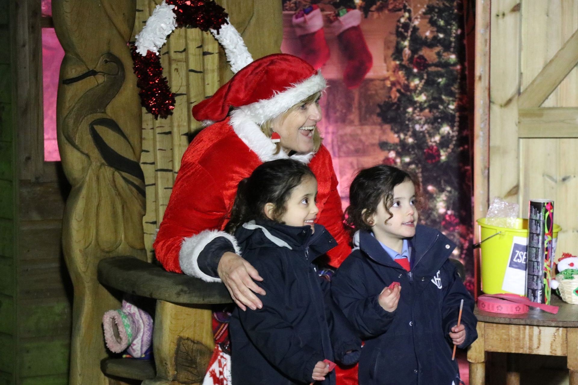 Woman in Santa suit with two children; festive decor, wooden backdrop.