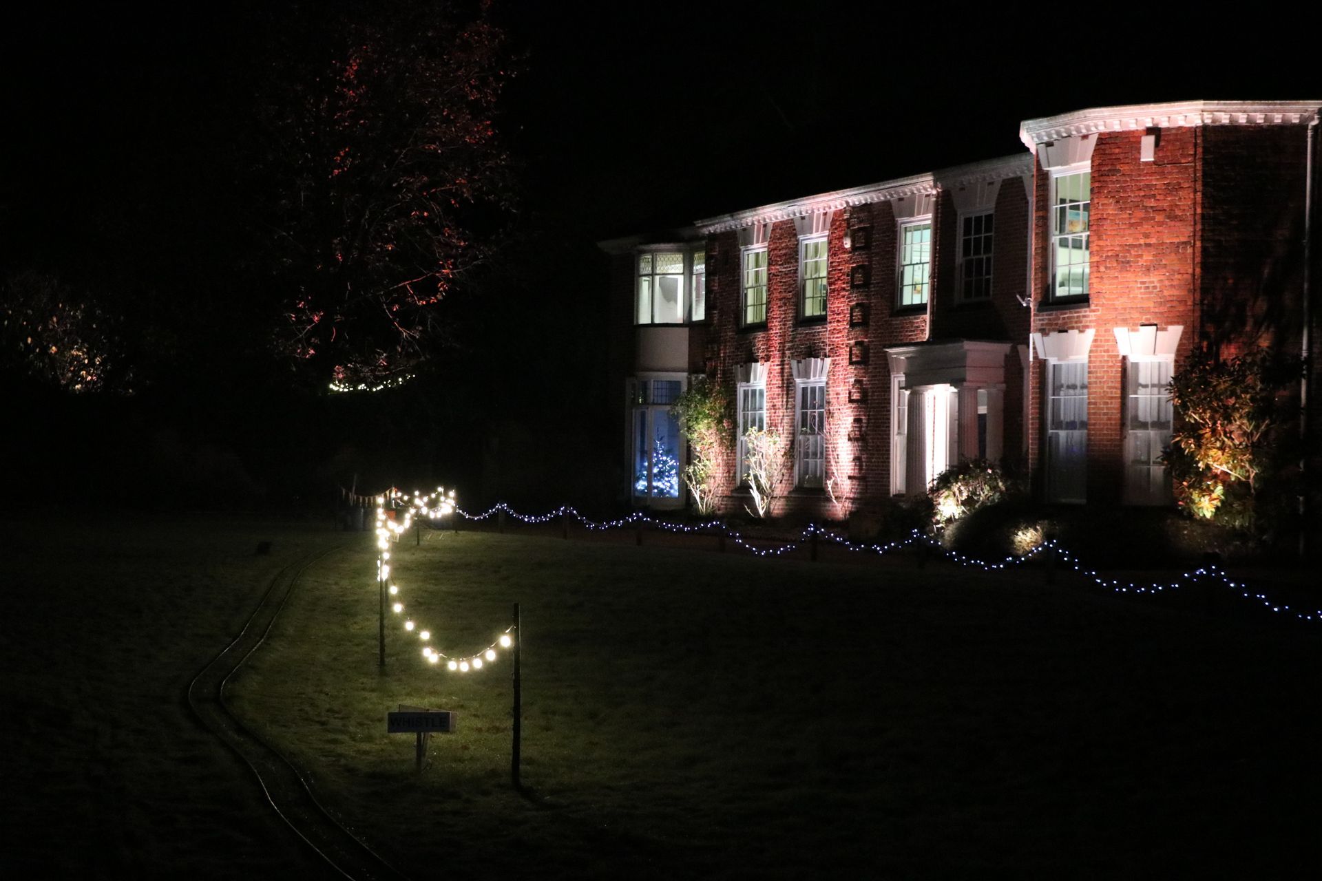 A brick house lit with white and red lights, front yard with string lights along the ground at night.