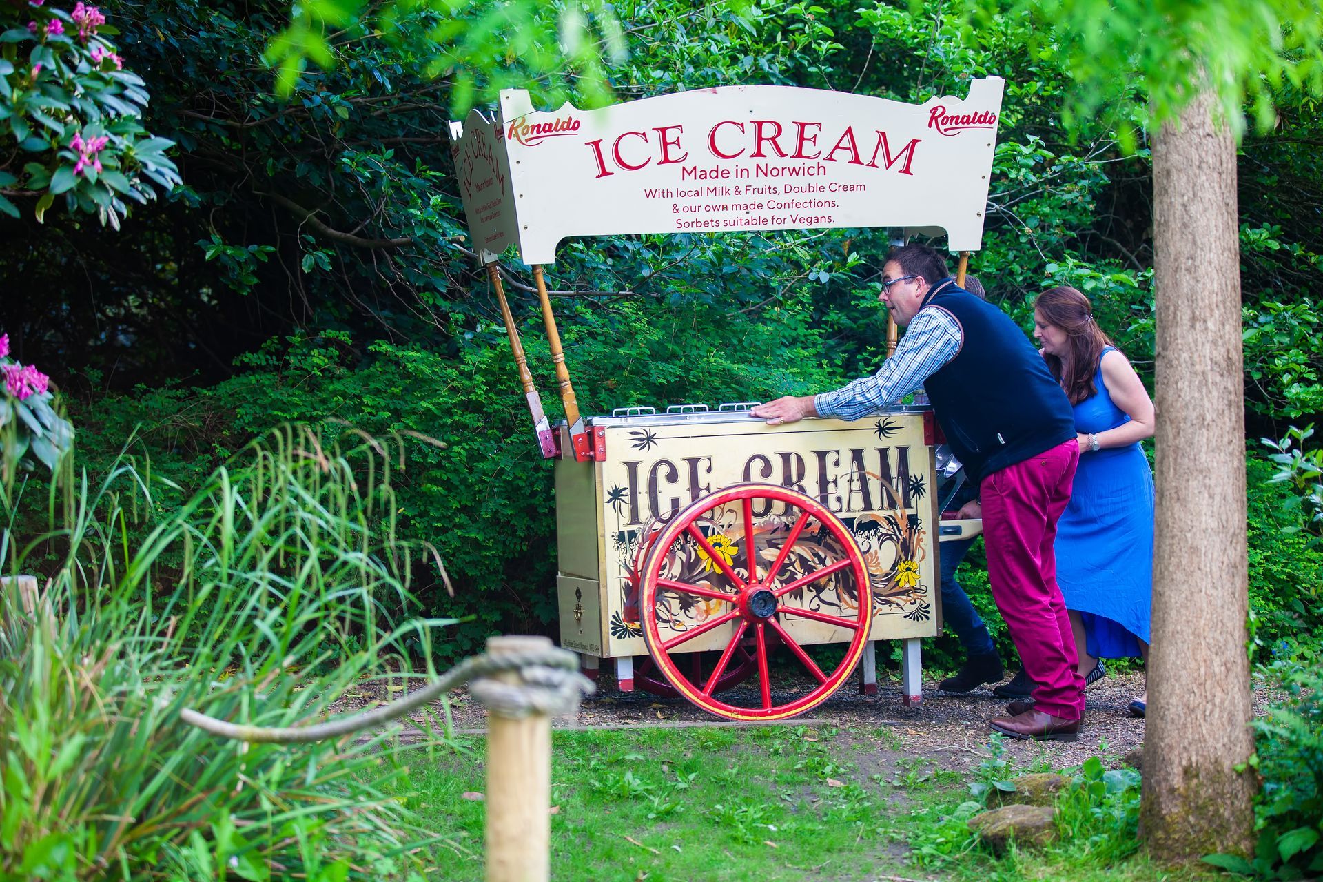 Couple by an antique ice cream cart in a garden, man pointing, woman smiling.