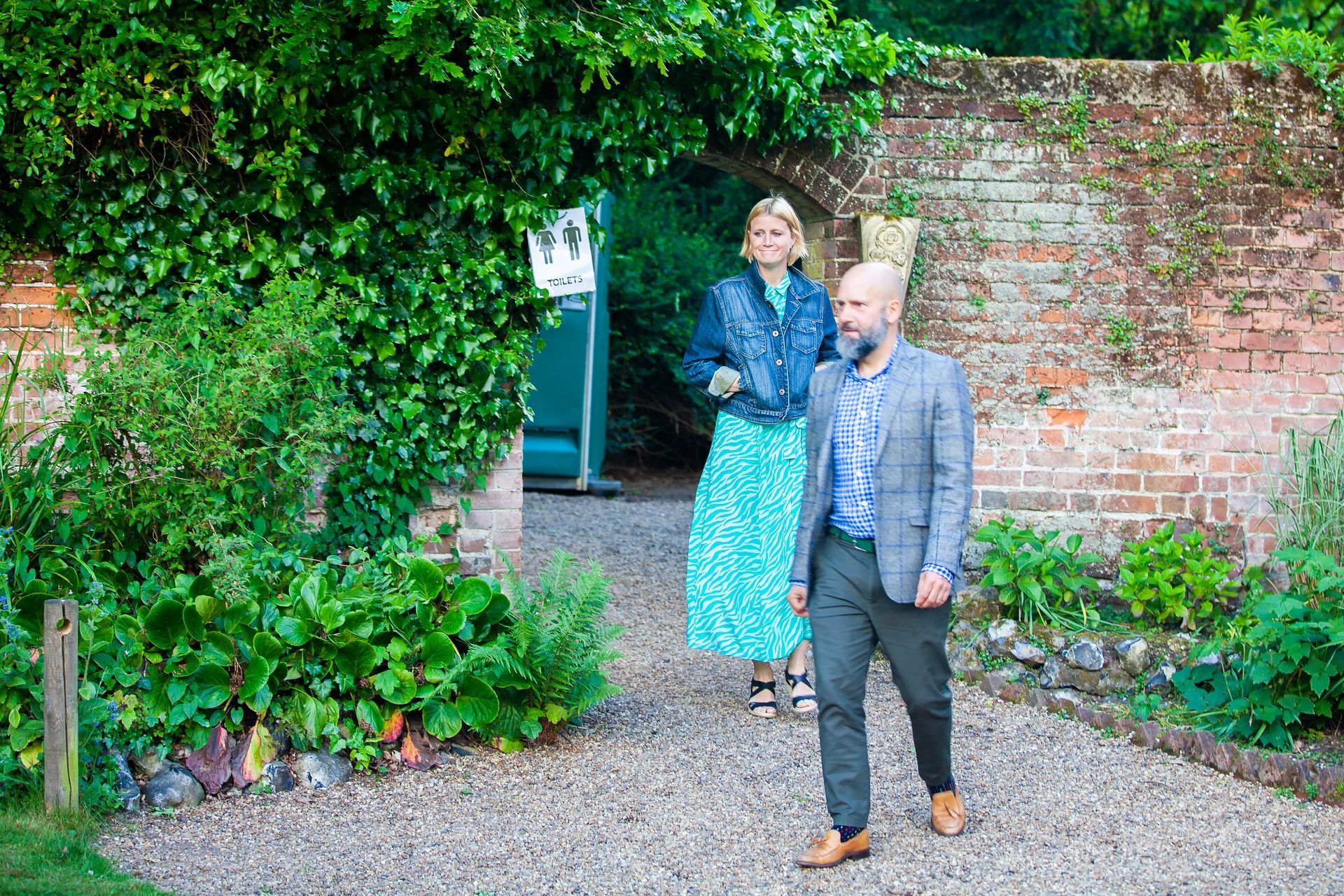 Woman and man walking on a gravel path. Man in suit. Woman in blue dress, jacket. Archway, greenery.