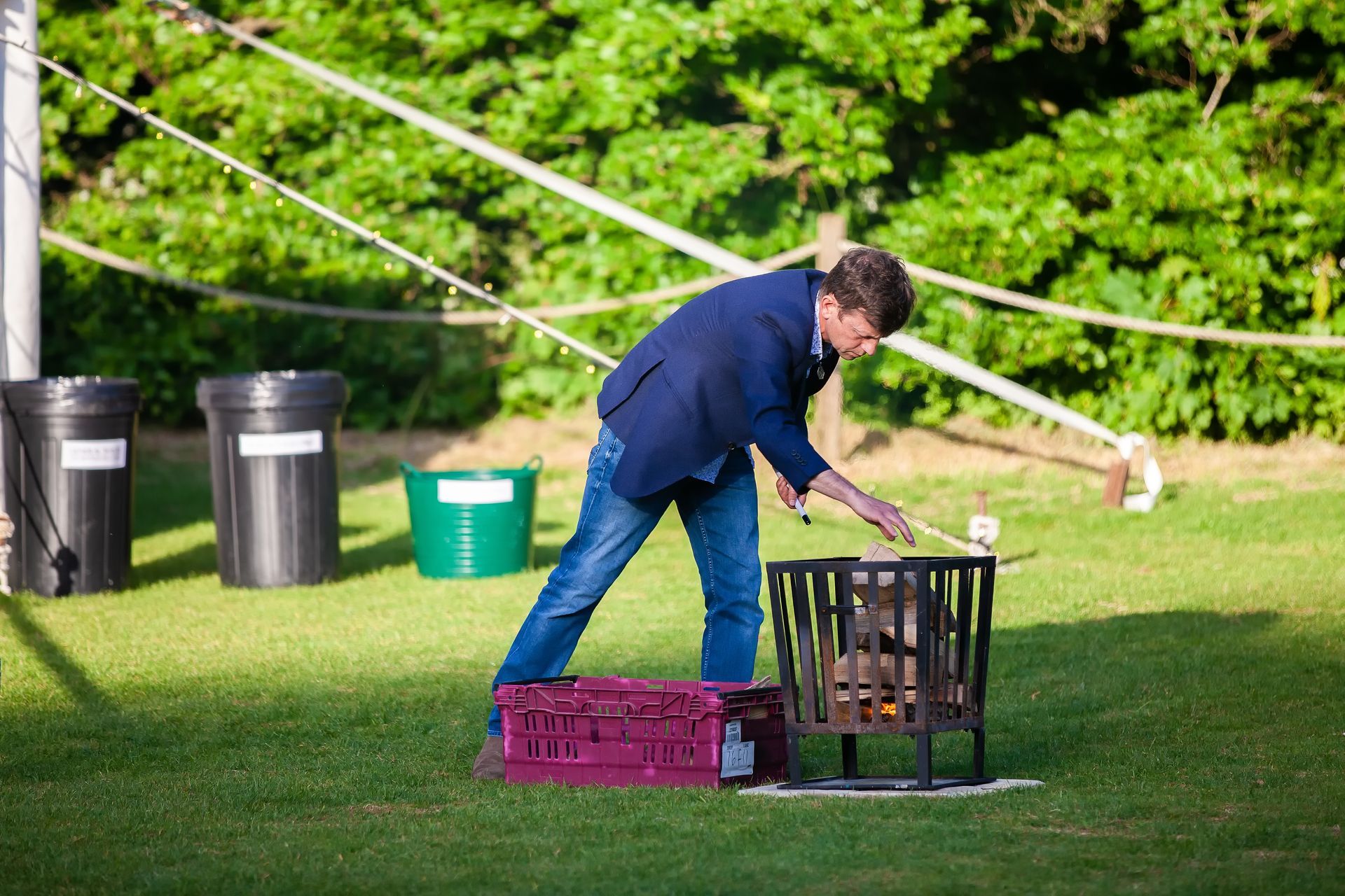 Man in blue blazer tends a fire pit on a green lawn, with trash cans and trees in background.