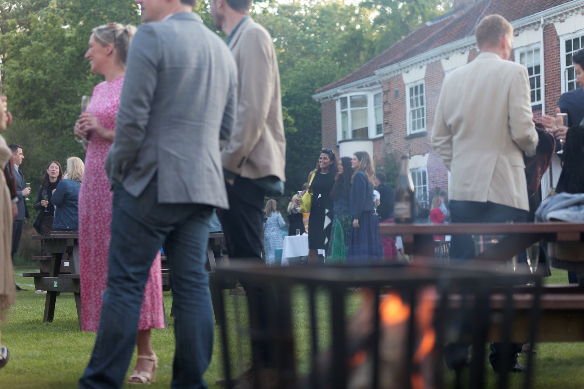 People socialize outdoors near a fire pit, house in background. Woman in pink dress, men in suits.