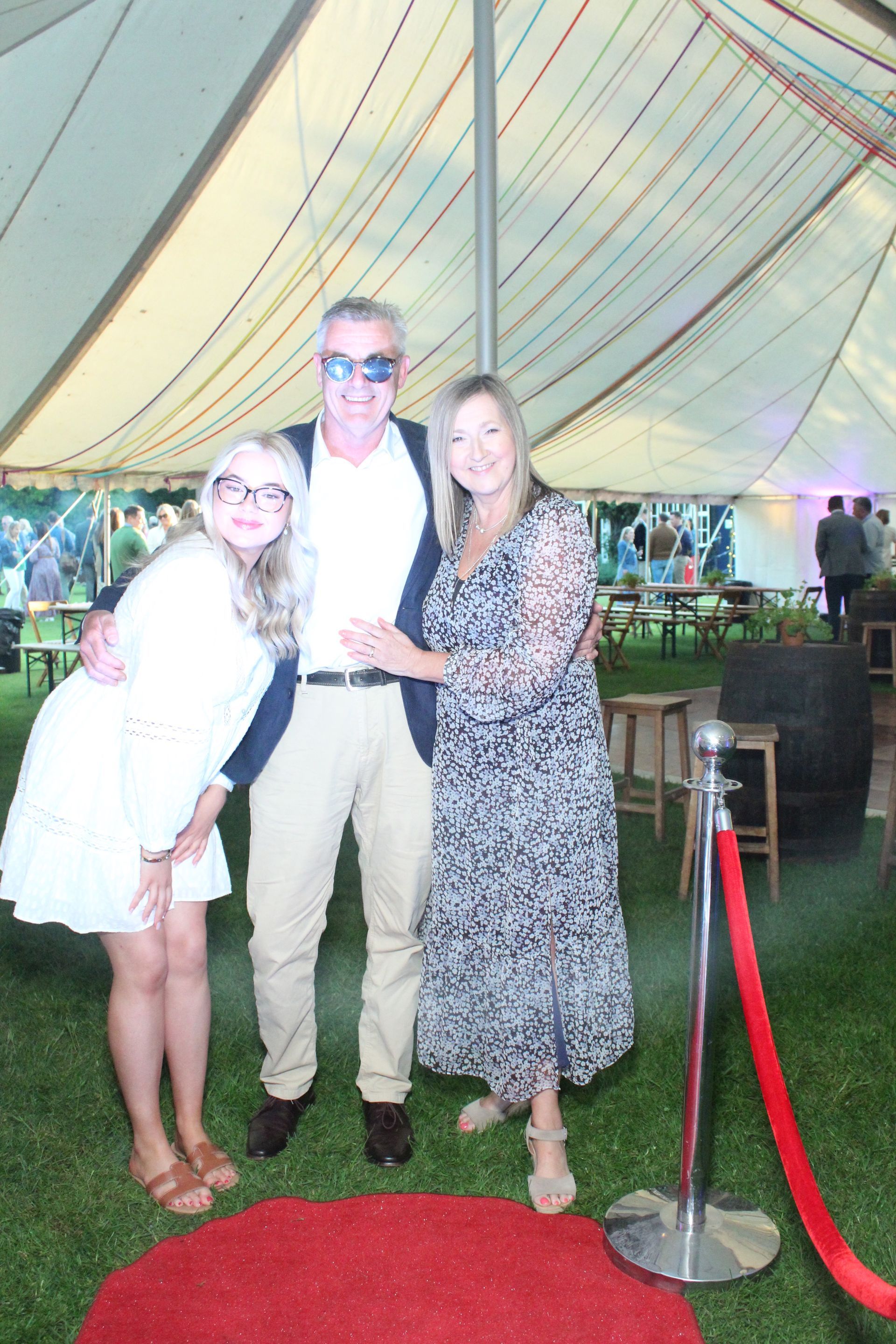 A family poses by a red carpet inside a tent. Woman in floral dress, man in sunglasses, and girl in white dress.