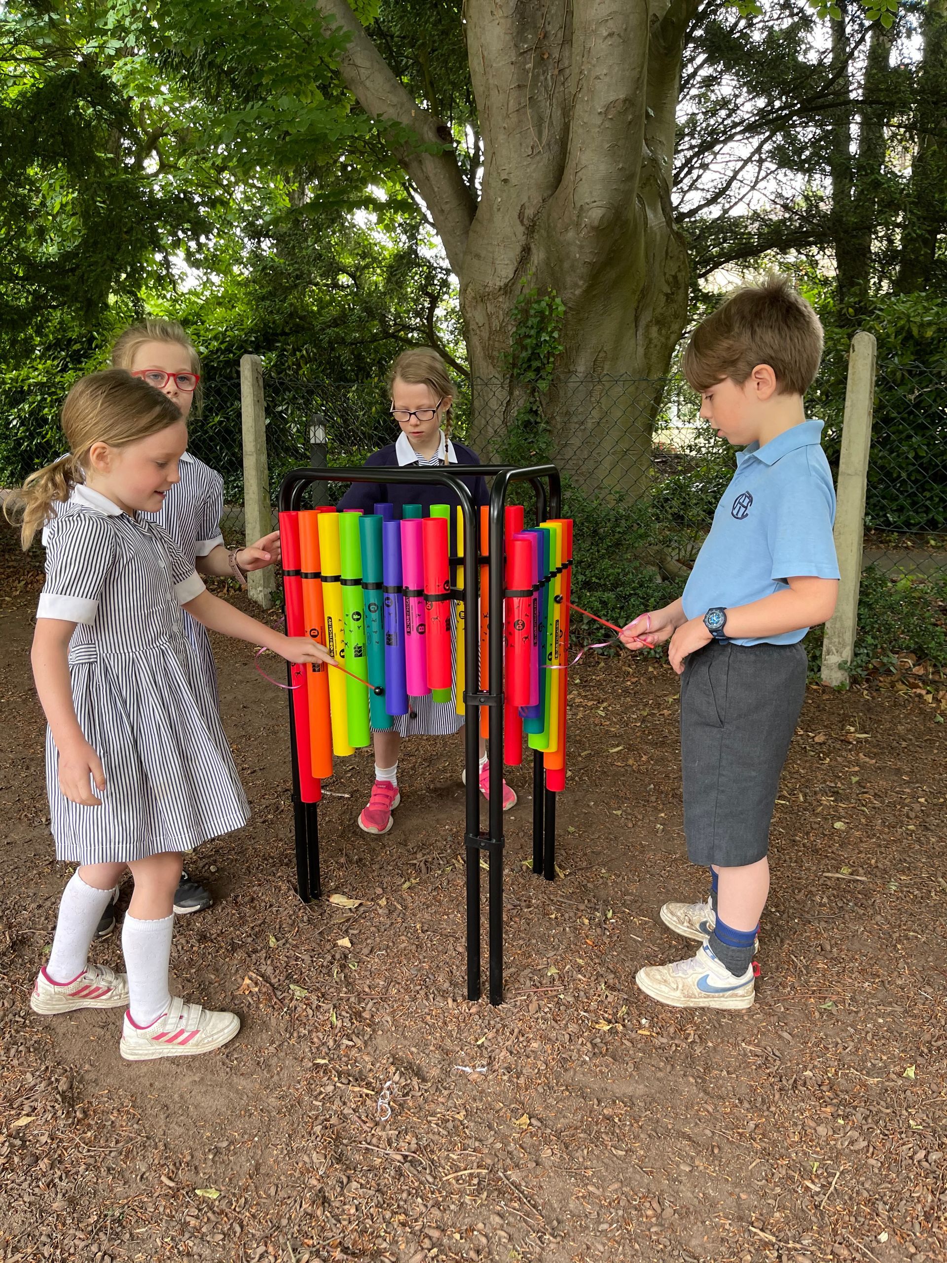 Four children playing a rainbow-colored outdoor xylophone in a wooded area.