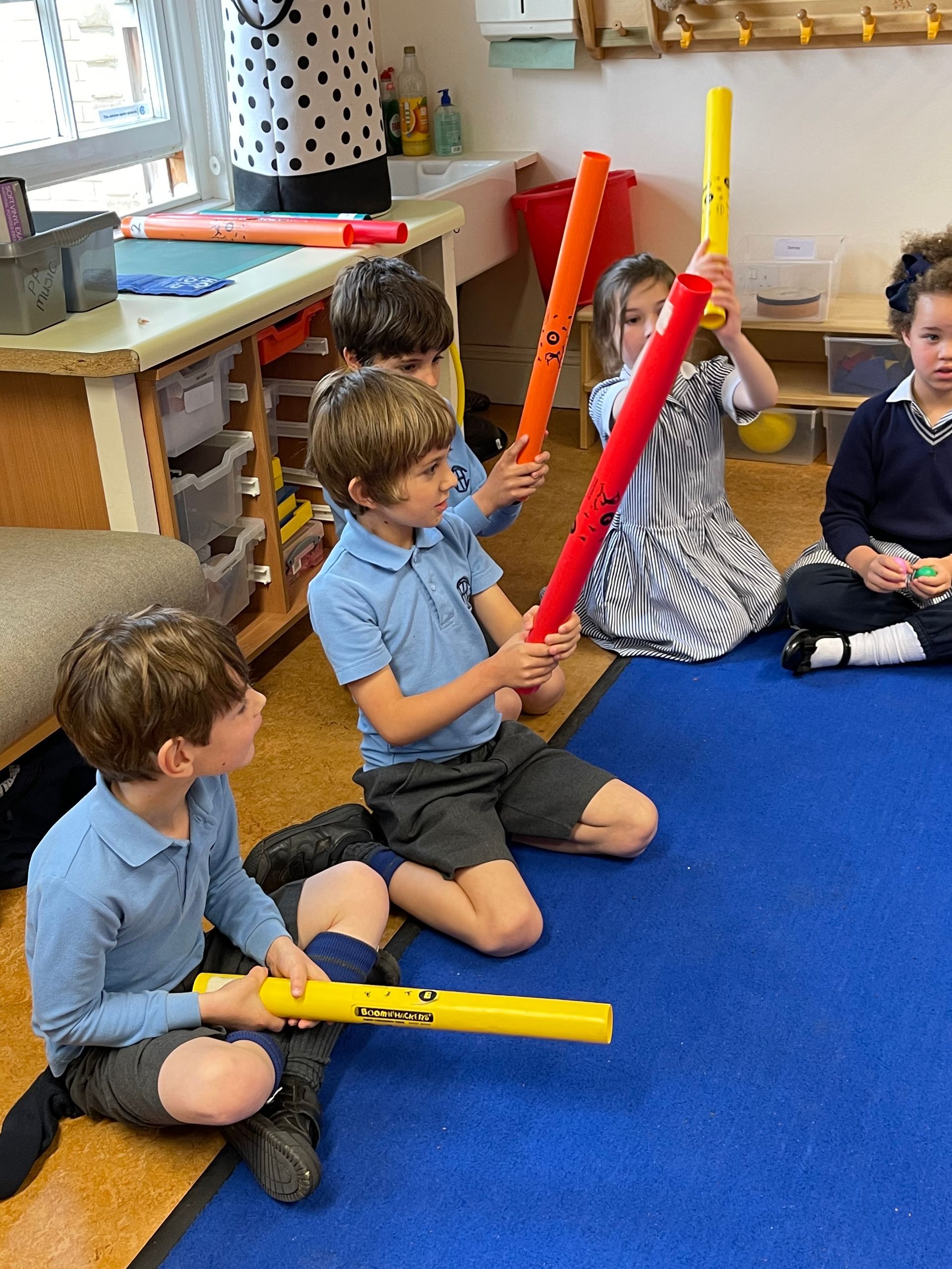 Children in a classroom playing musical instruments.  Some are kneeling on a blue mat and holding colorful tubes.