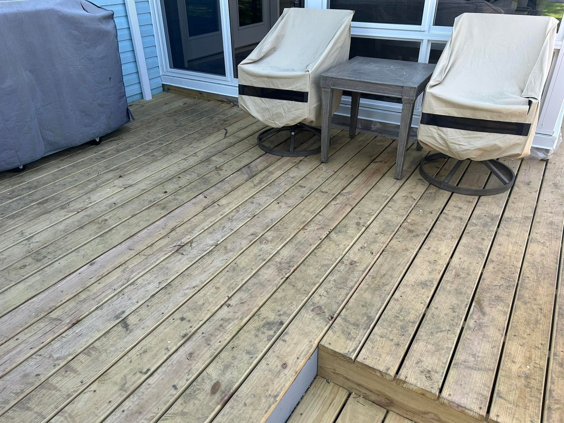 Wooden deck with two beige lounge chairs, a small table, and a covered grill beside sliding glass doors