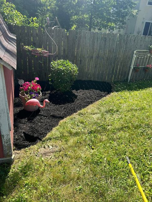 Backyard garden bed with dark mulch, pink flowers, a fence, and sunlit grass slope