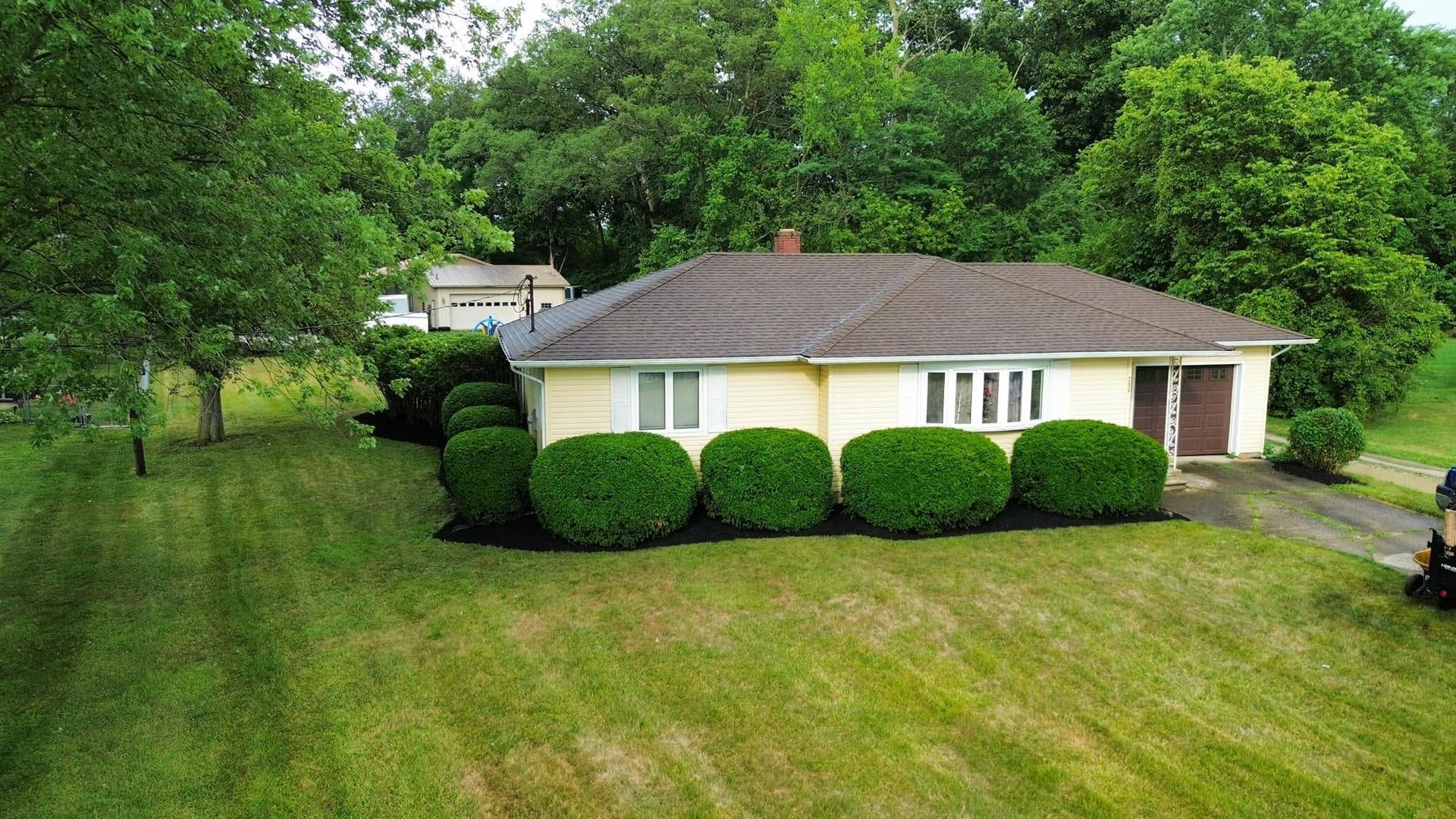 Single-story beige house with a brown roof, trimmed bushes, and a green lawn under trees