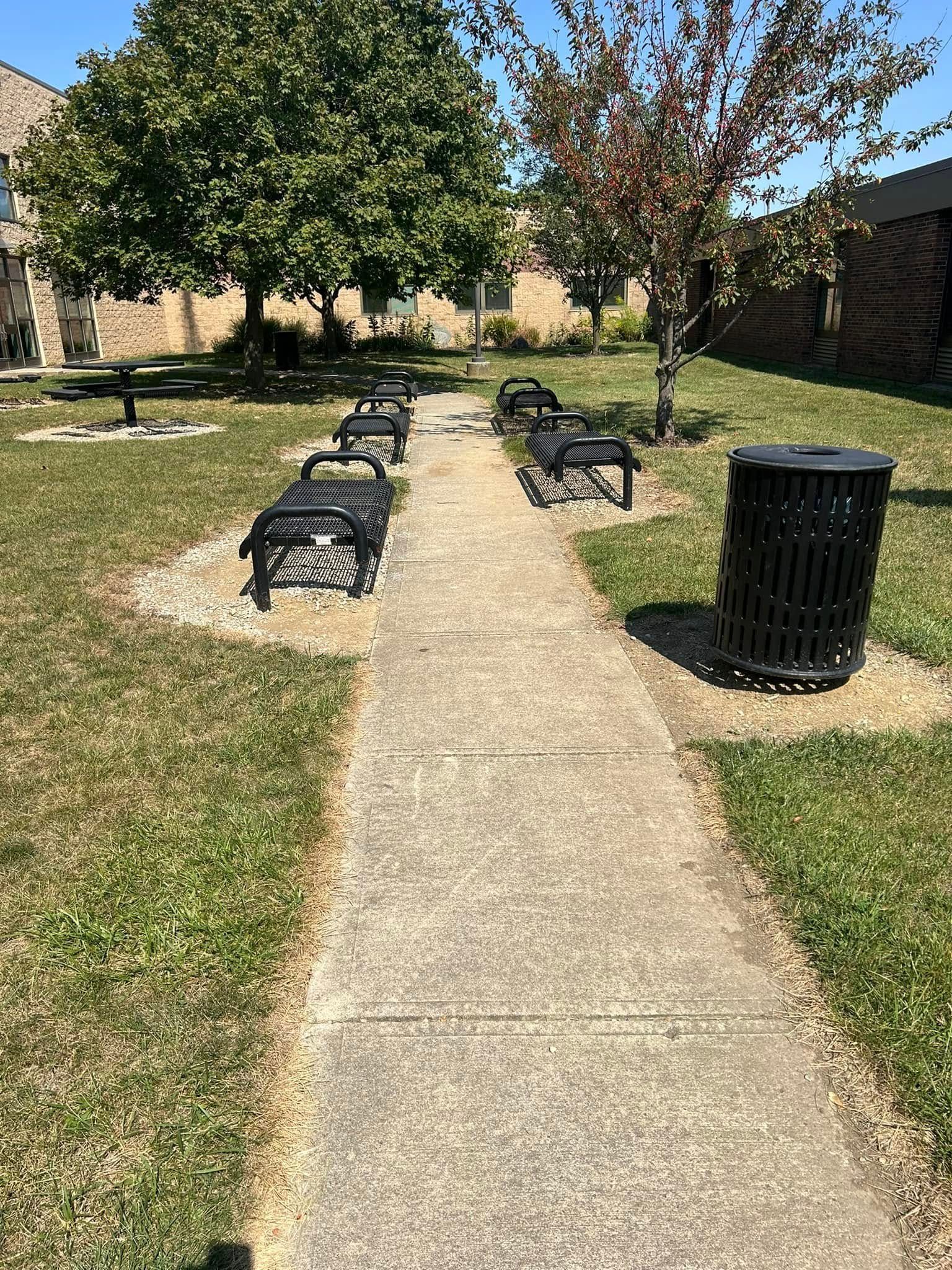 Concrete path between black benches and a trash can in a sunny courtyard with trees and grass