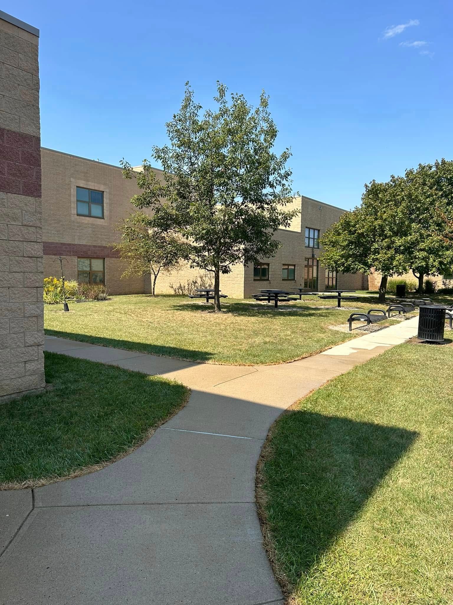 Courtyard with concrete walkway, grass, trees, and a brick building under a sunny sky