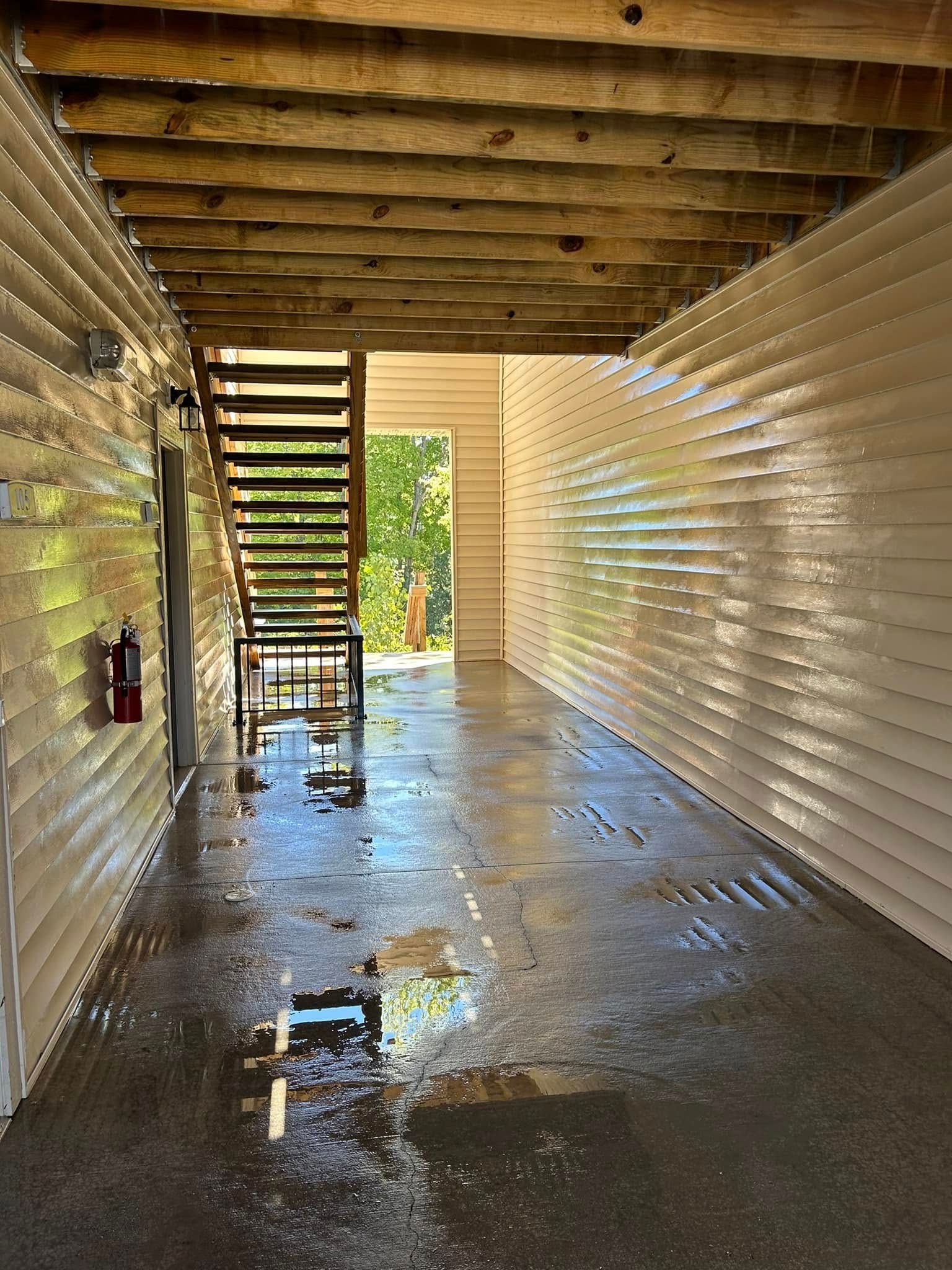 Wet corridor under a wooden staircase, with corrugated metal walls and bright sunlight at the far end.