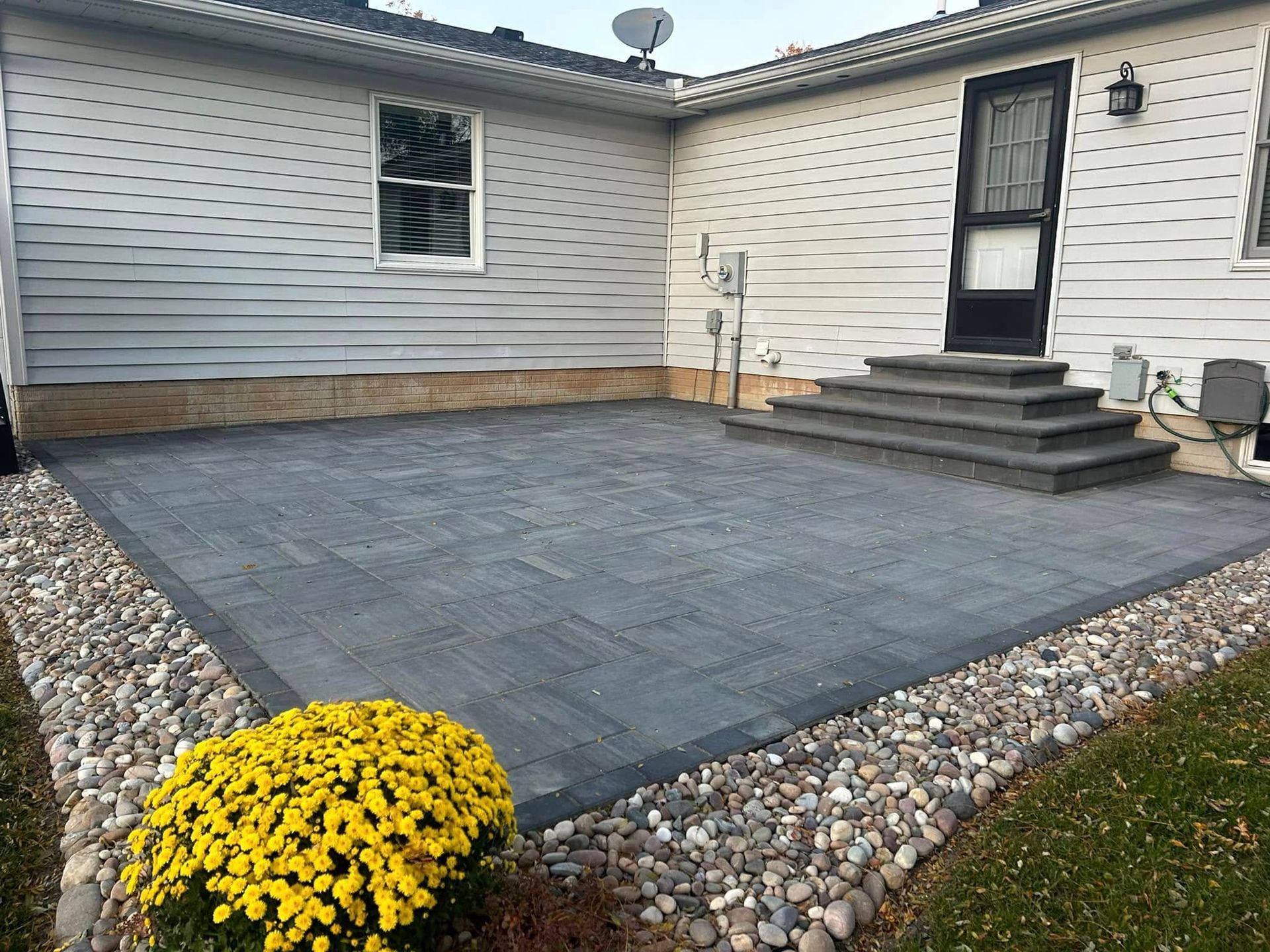 Backyard patio with gray pavers, white siding, steps, and a yellow flower bed in front
