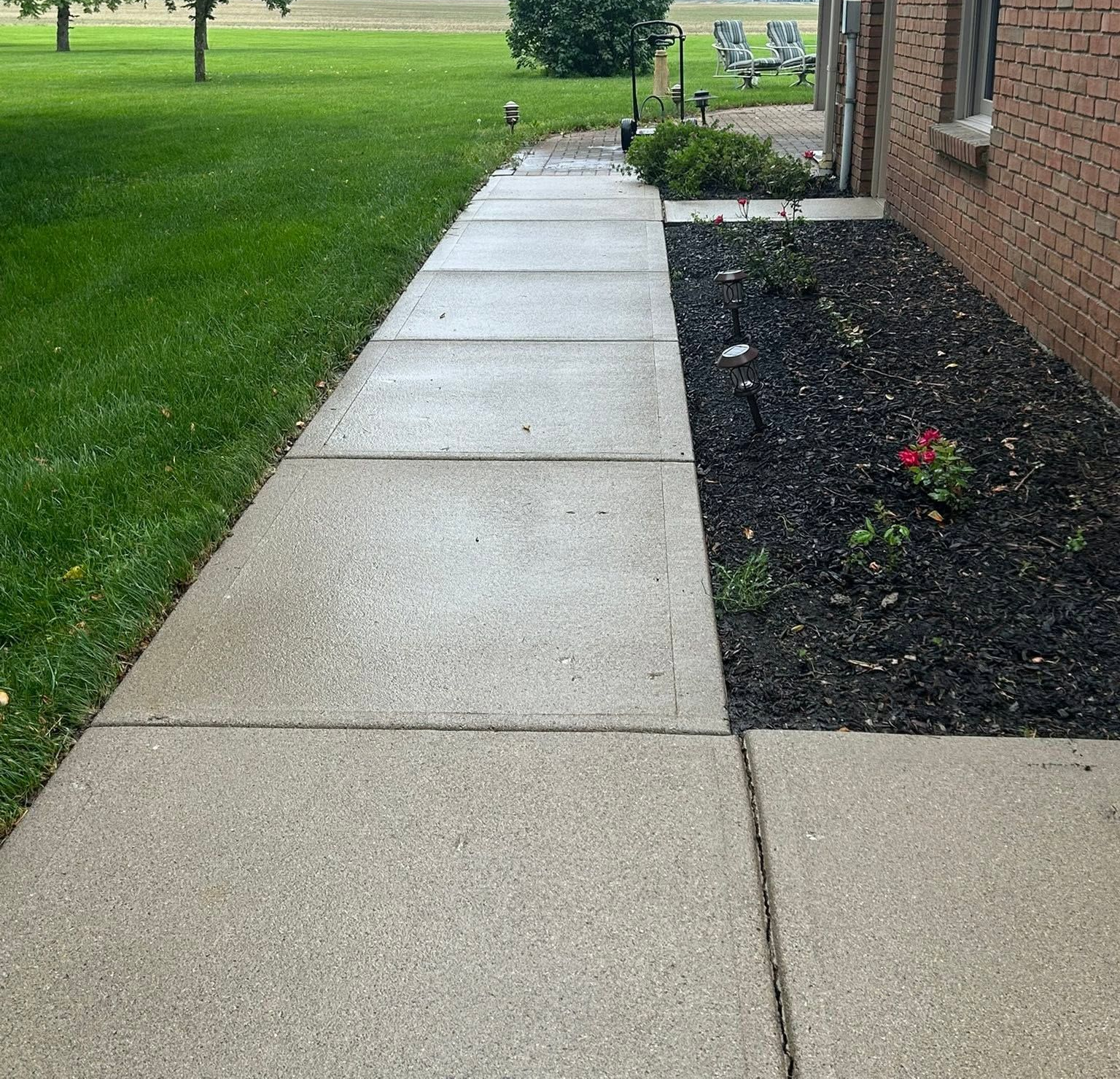 Concrete sidewalk beside a brick building, with green lawn and flower beds along the path.