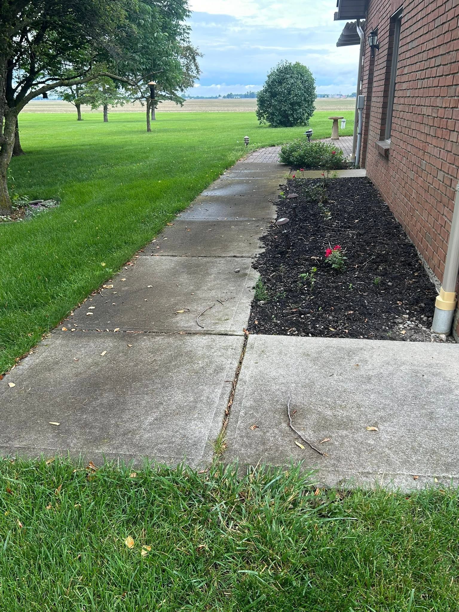 Concrete walkway beside a brick house and grassy yard, leading toward open fields.