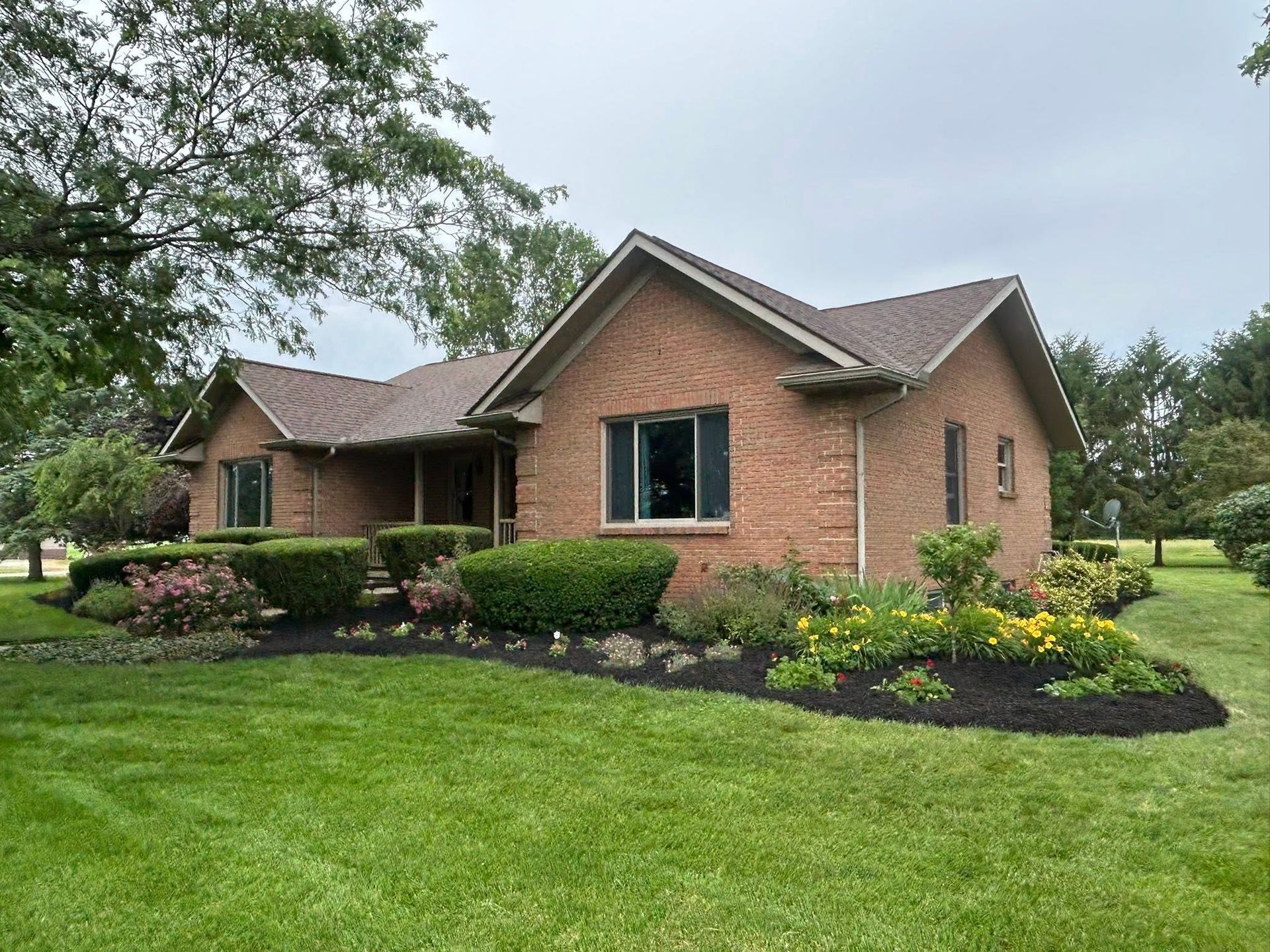 Brick suburban house with manicured lawn, shrubs, and landscaped flower beds under an overcast sky