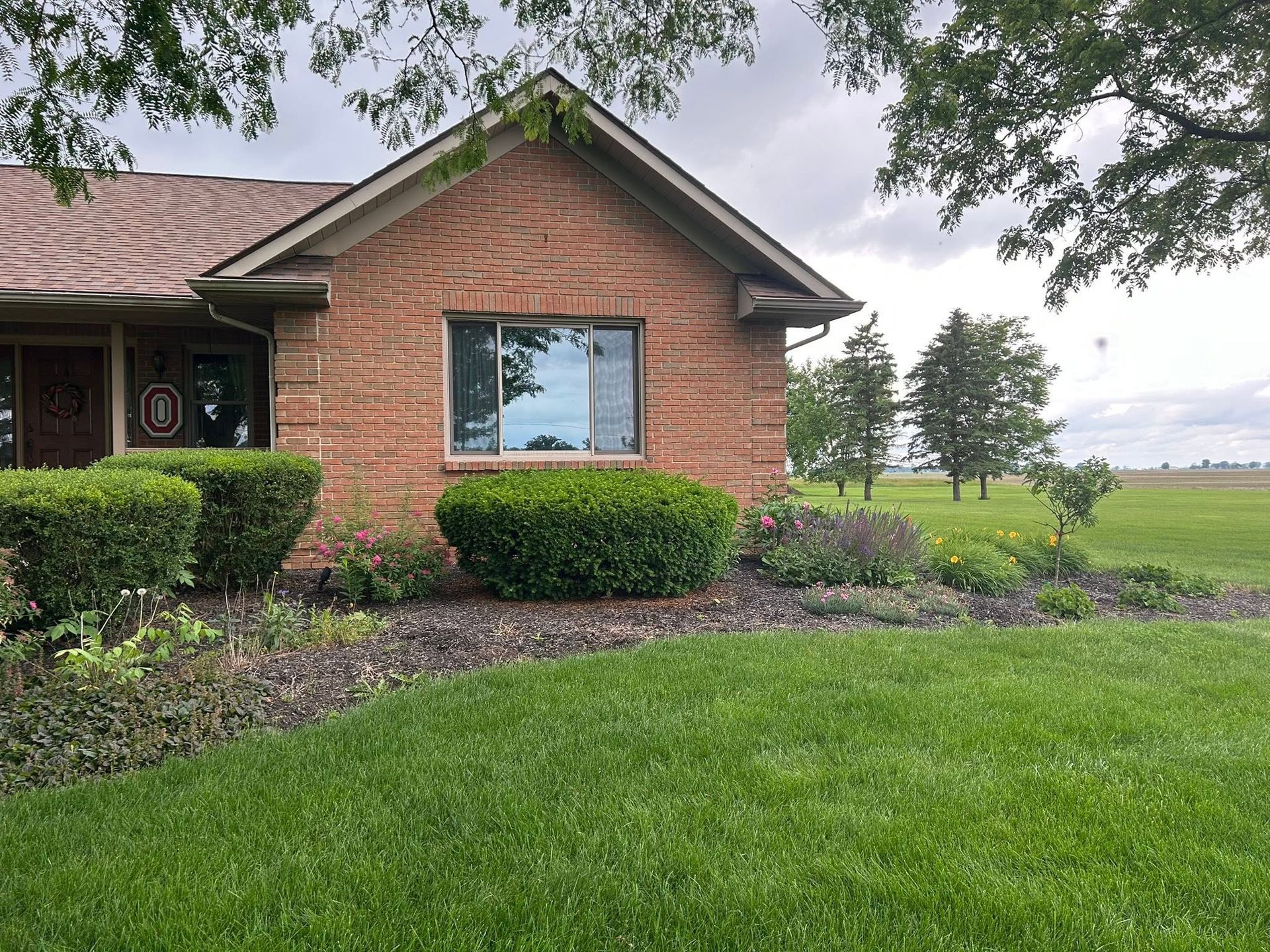 Brick house with trimmed shrubs and a green lawn beside open fields under cloudy skies