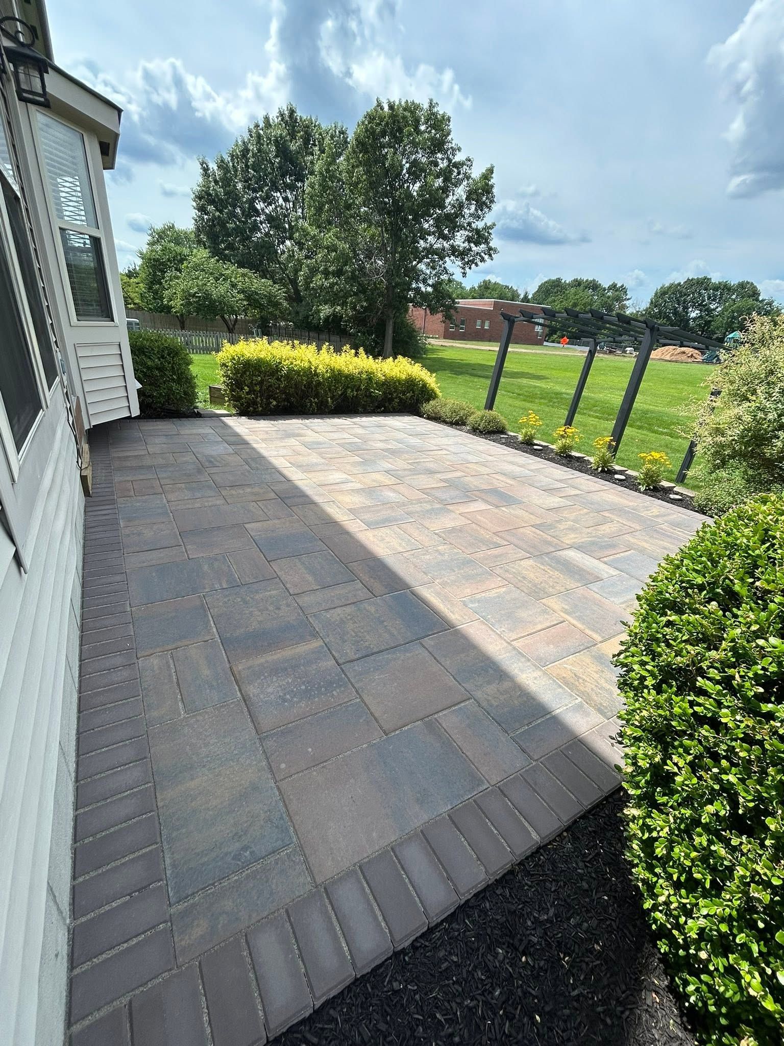 Back patio with gray pavers, yellow shrubs, and a grassy yard under a bright sky