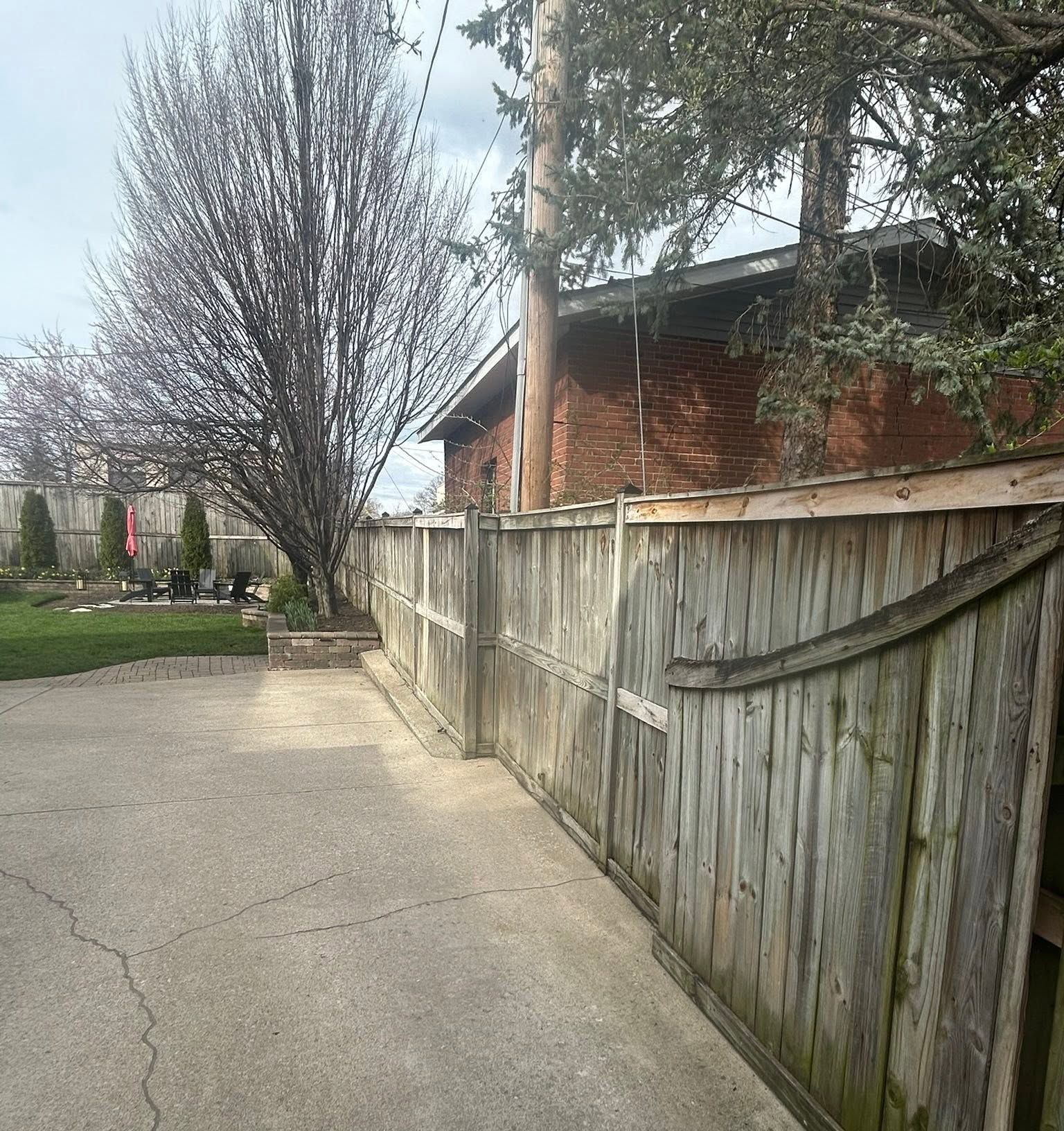 Concrete driveway beside a weathered wooden fence, with a red-brick house and leafless tree