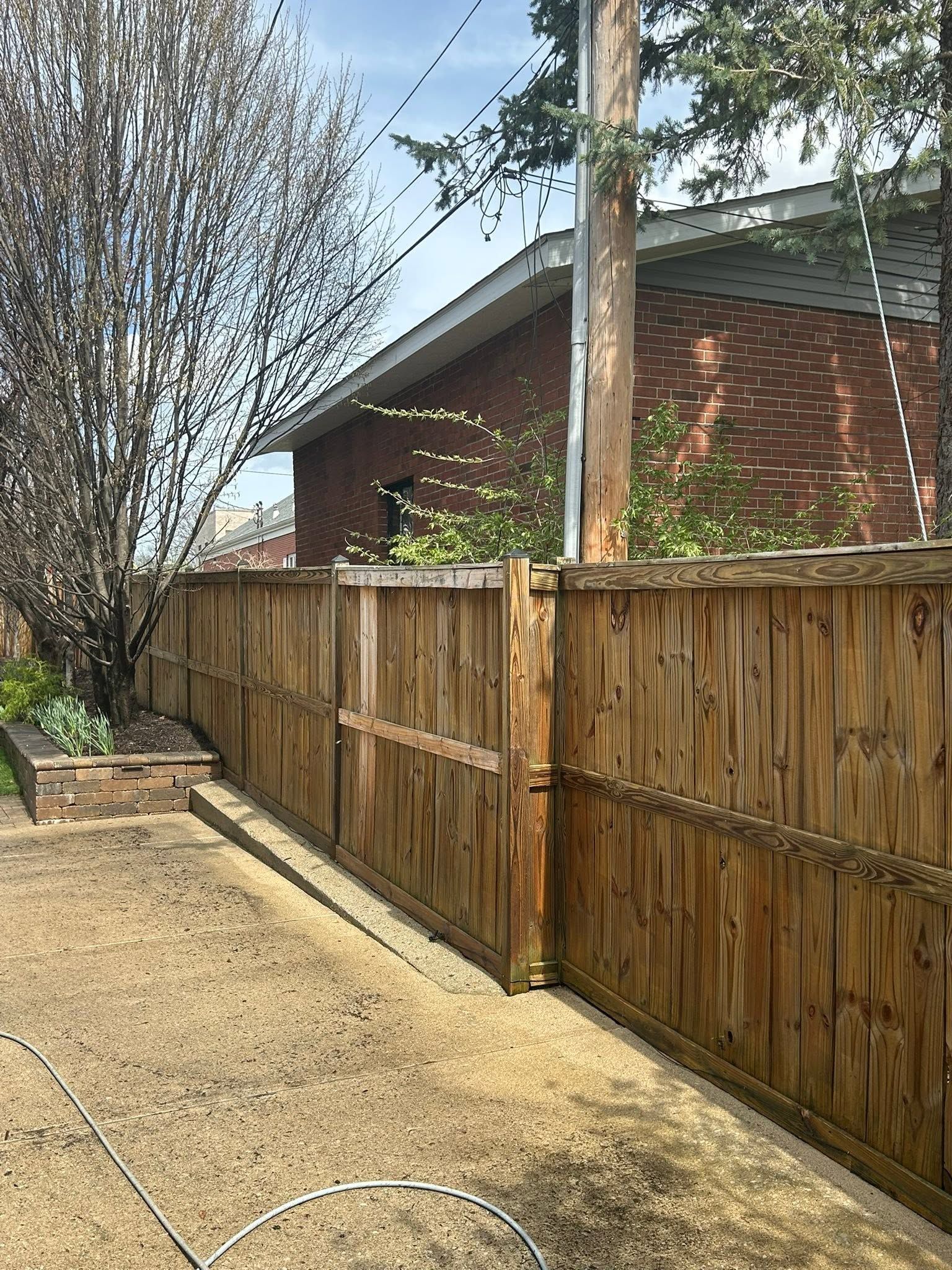 Wooden fence beside a gravel driveway, with trees and a brick house under a blue sky.