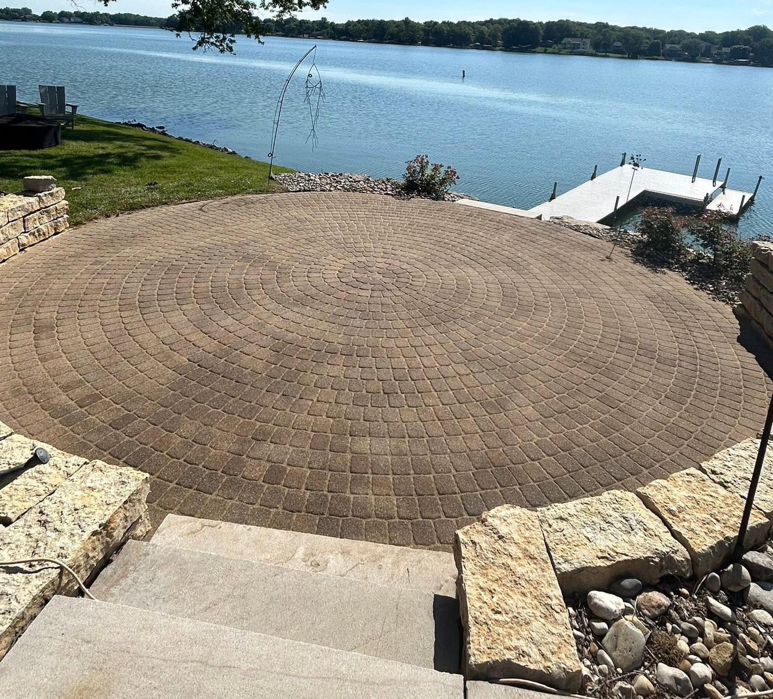 Circular brick patio by a lake with steps and a dock under a clear sky