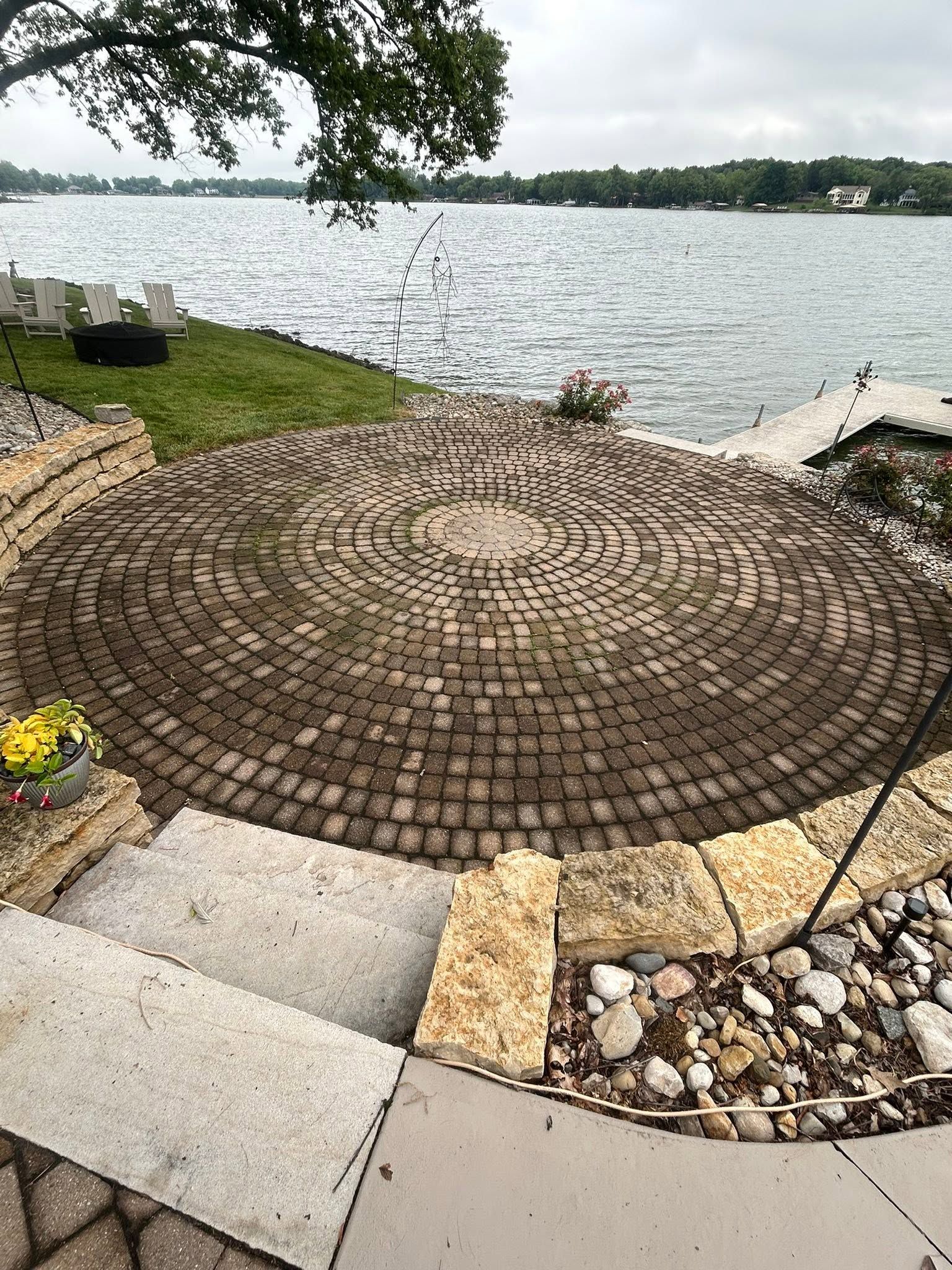 Stone patio by a lakeshore with chairs, steps, and flowerpots; water and trees in the background