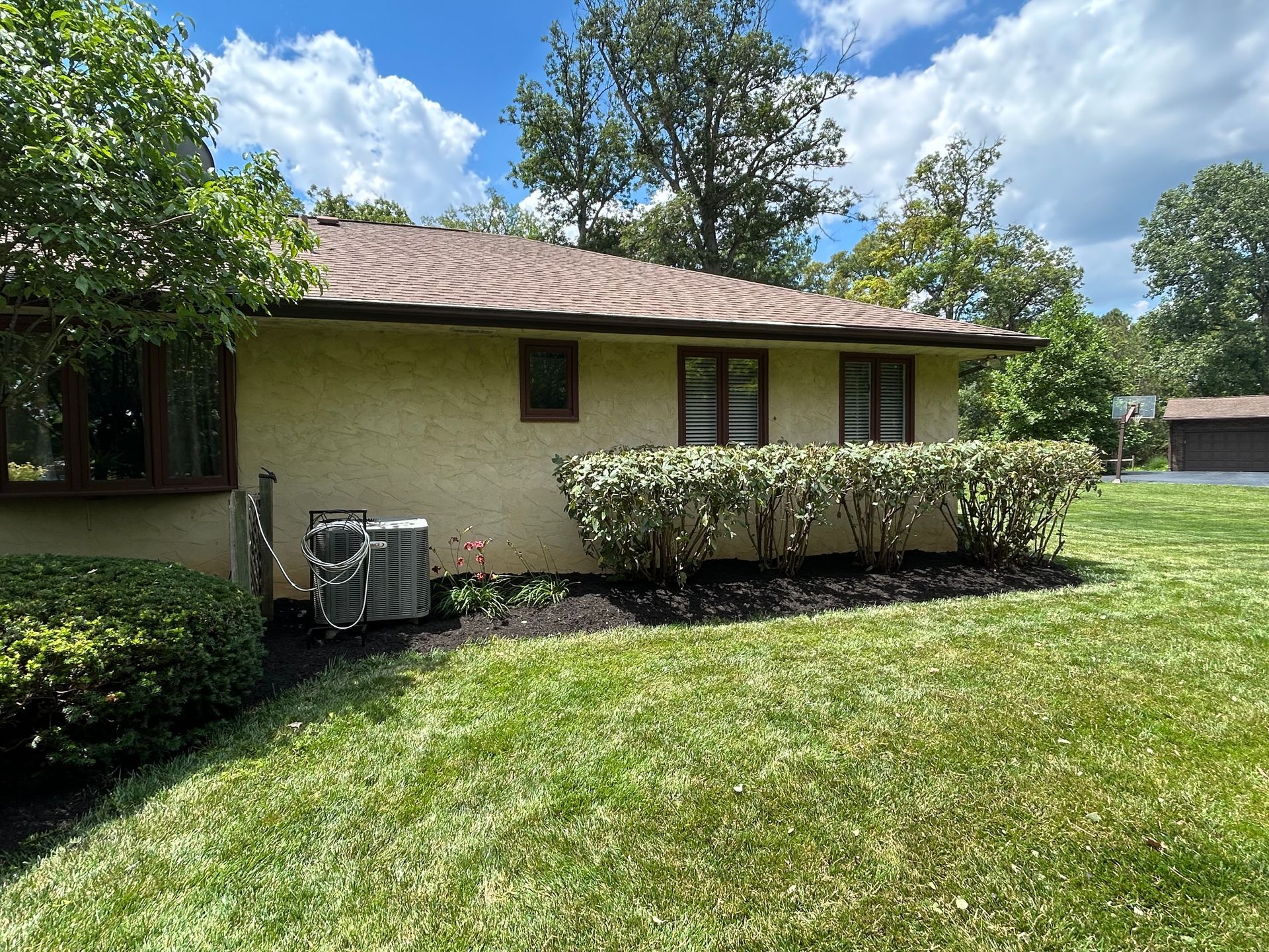 Single-story beige house with a green lawn, trimmed shrubs, and trees under a blue sky.