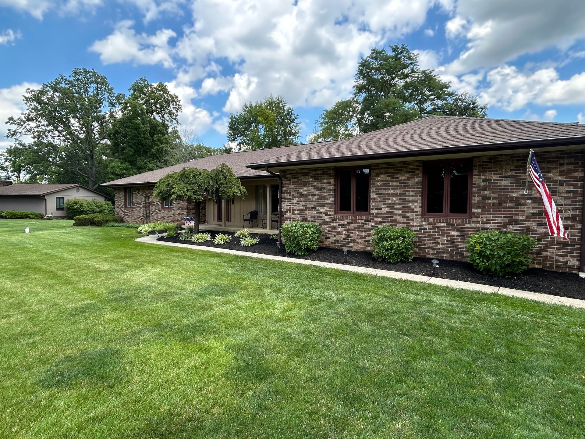 Single-story brick house with a manicured lawn, trees, and an American flag by the front porch